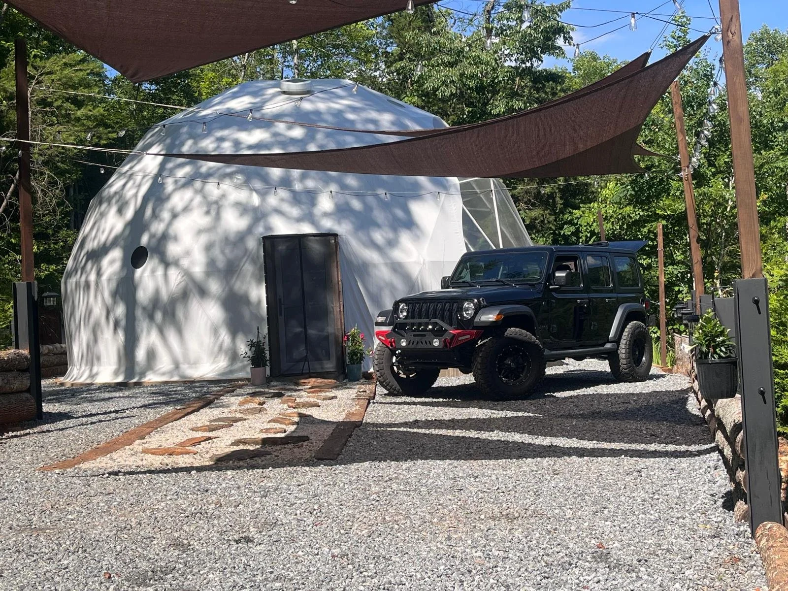 A modern dome-shaped white tent, a black Jeep with off-road tires, and a gravel pathway with a stone border. There are brown shade sails overhead and lush green trees in the background.