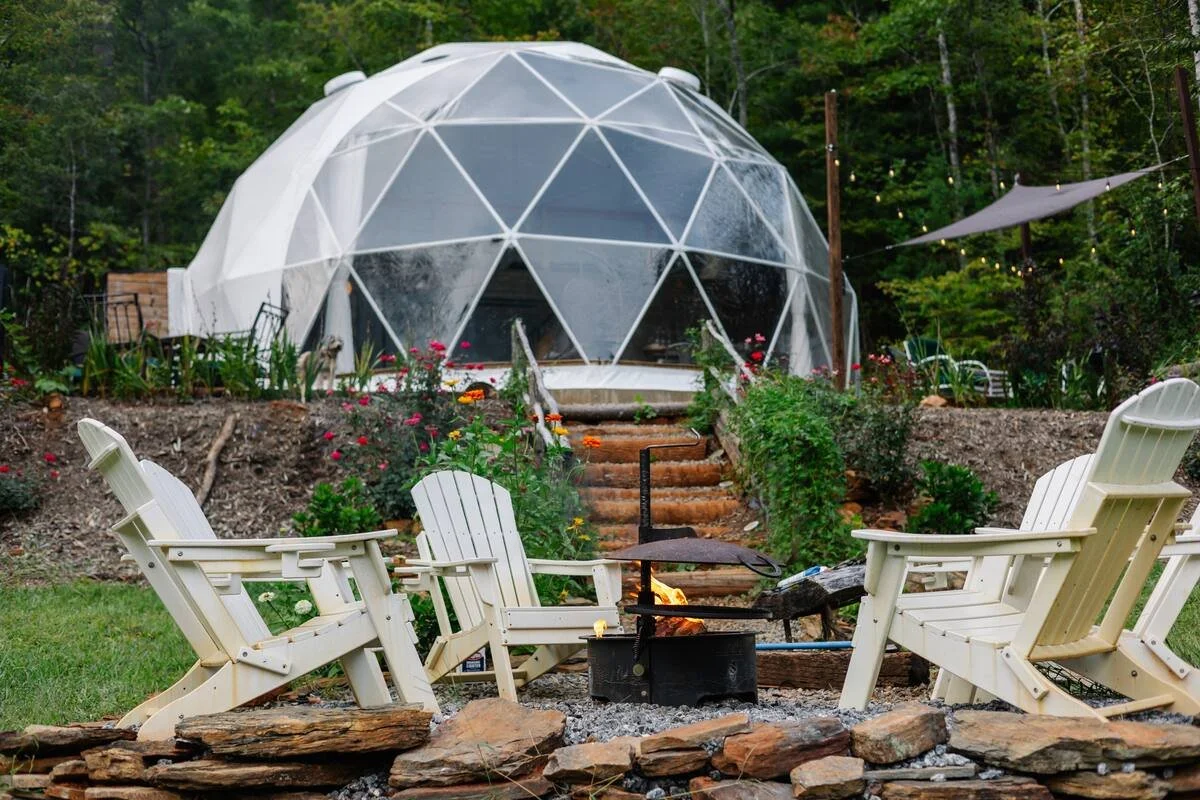 A backyard scene featuring four white Adirondack chairs arranged around a fire pit, with a garden and steps leading up to a geodesic dome tent in the background, surrounded by trees and greenery.