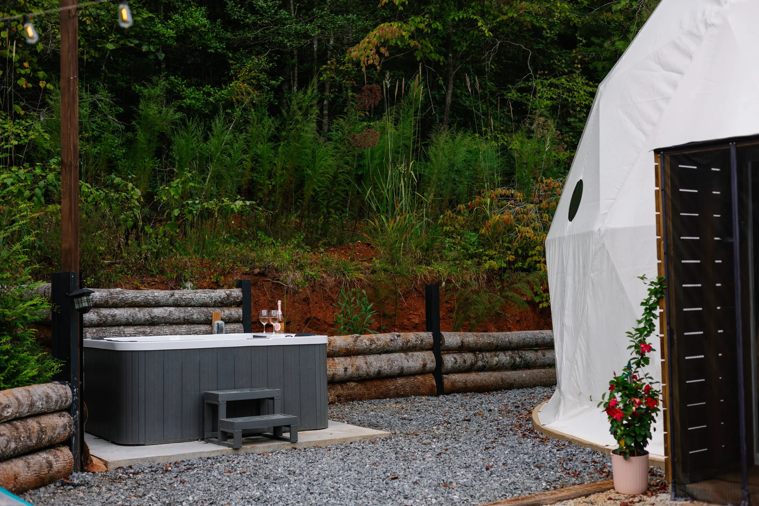 Outdoor scene with a hot tub on a gravel surface, wine glasses on the edge, a small step stool, and a white tent with a potted plant nearby, surrounded by green trees and a wooden log fence.