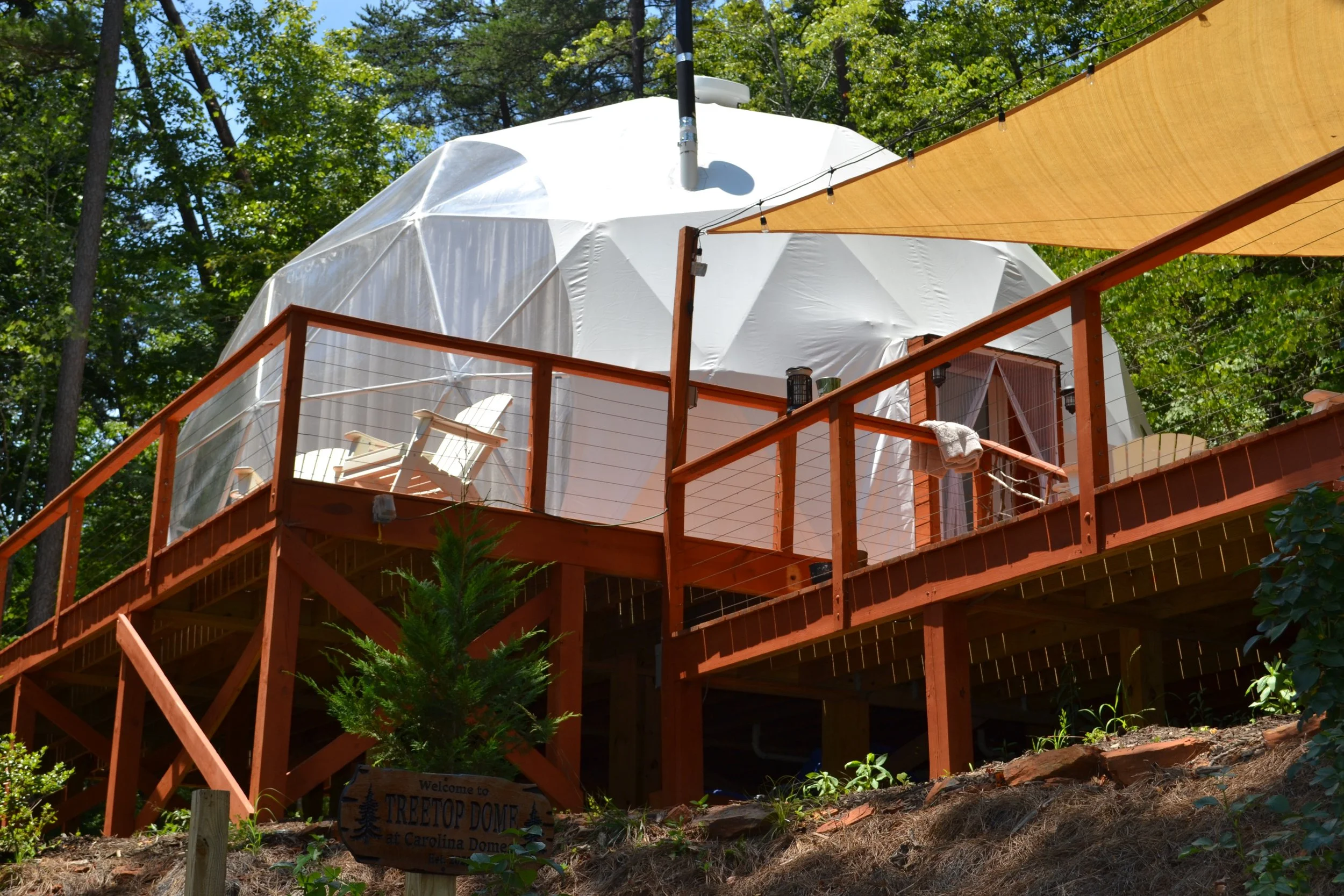 A wooden deck with chairs and a towel, supporting a large dome-shaped structure with a white fabric cover, surrounded by green trees.