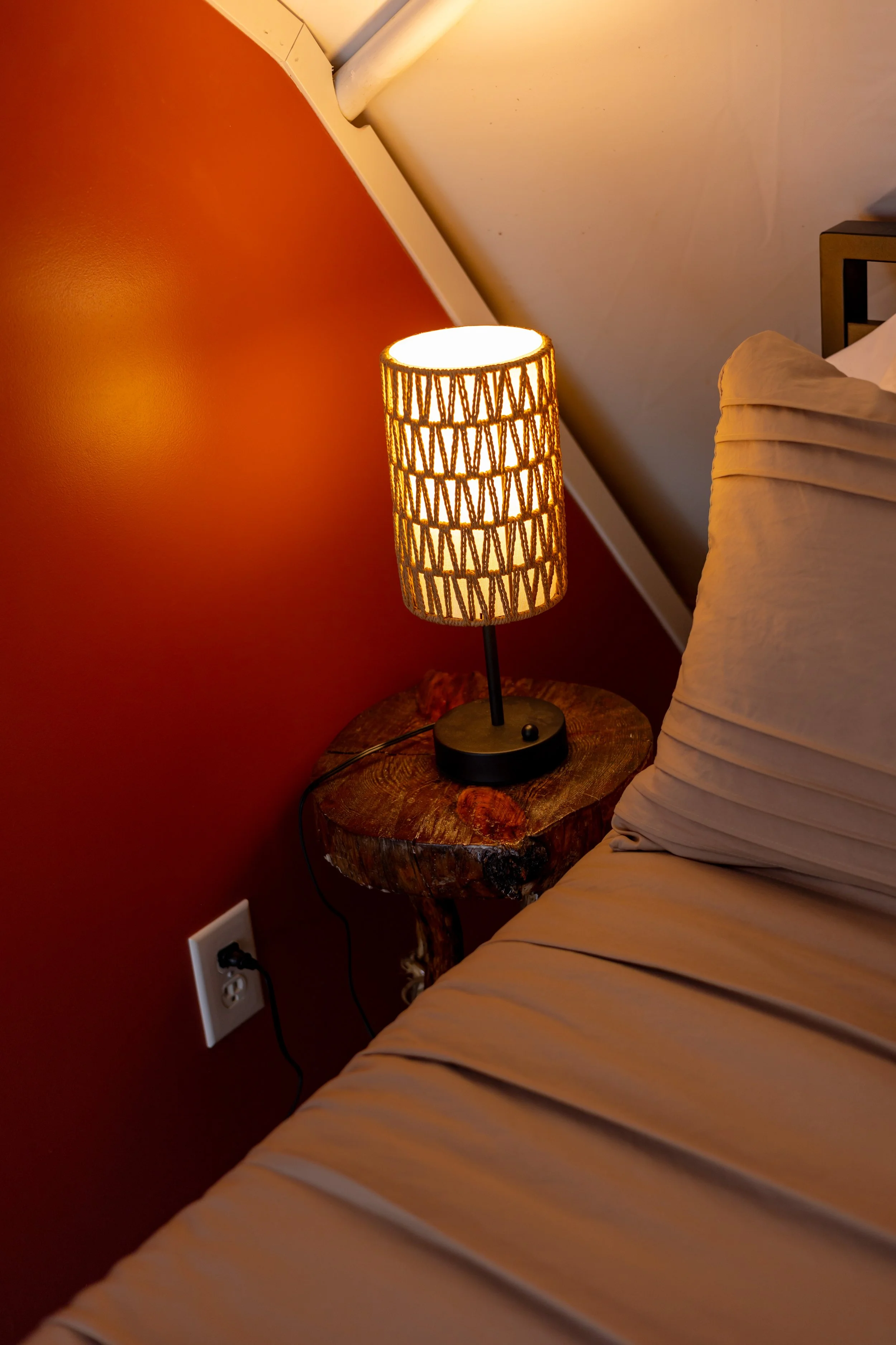 A bedside lamp with a woven lampshade on a rustic wooden nightstand next to a bed with beige sheets in an angled corner of a room with a red wall and sloped ceiling.
