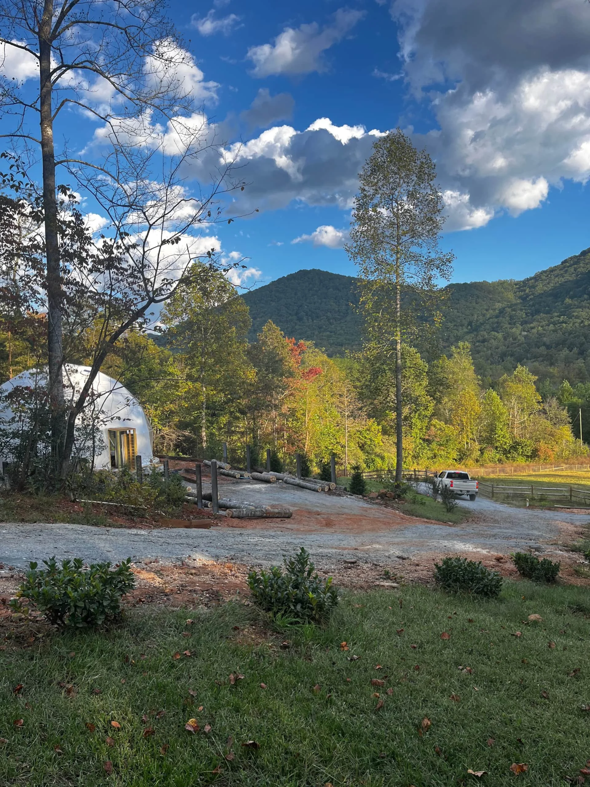 A scenic outdoor scene with a gravel driveway, a white truck, and a white geodesic dome structure surrounded by trees with green and some fall-colored leaves, mountains in the background, and a dramatic partly cloudy sky.