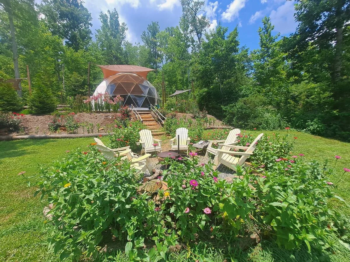 A lush garden with colorful flowers surrounds a circle of white Adirondack chairs, with a geodesic dome structure and trees in the background under a partly cloudy sky.