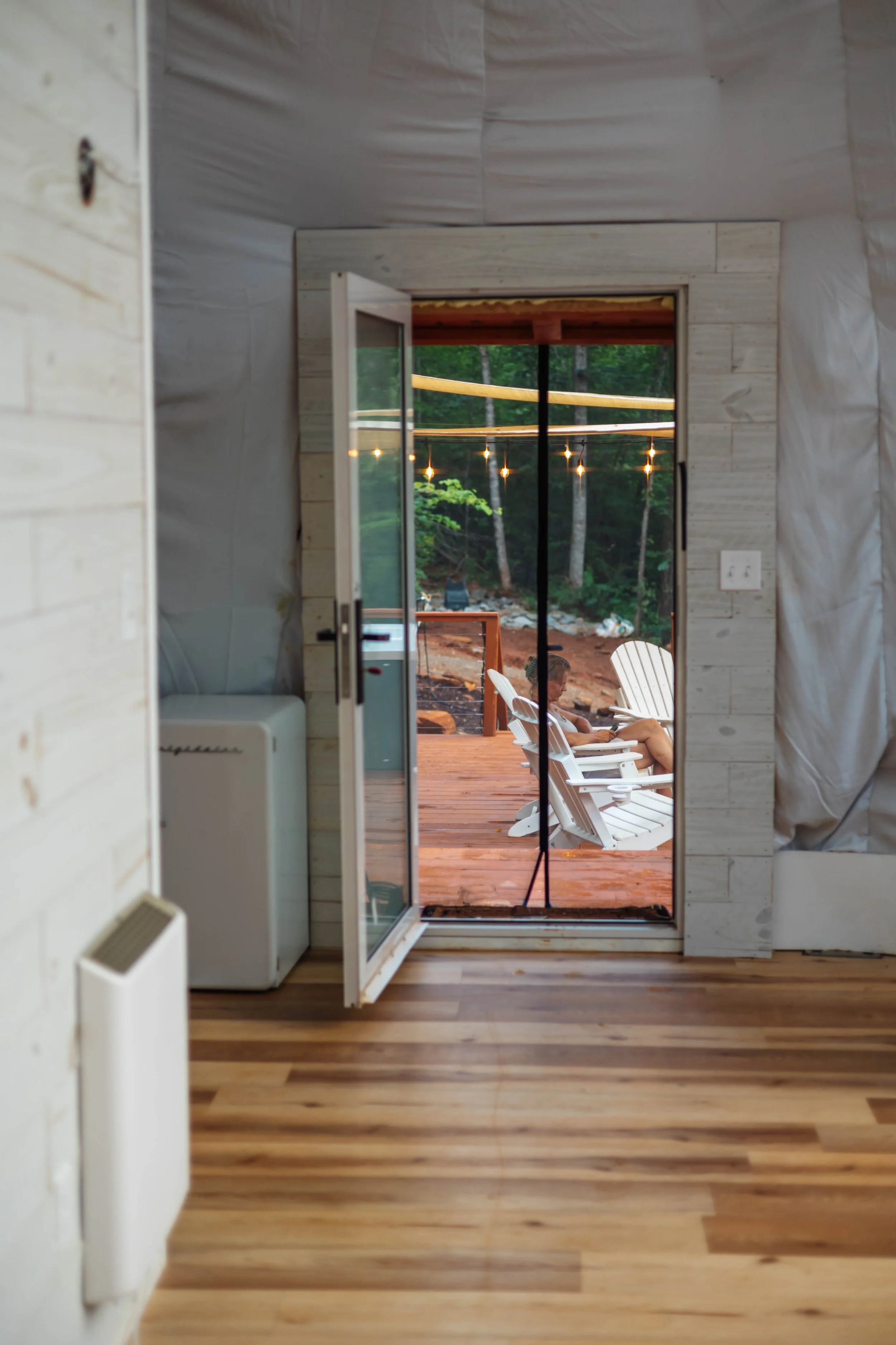 View through open glass door to a wooden deck with two white Adirondack chairs, a child sitting on one, overlooking a wooded outdoor area with string lights.