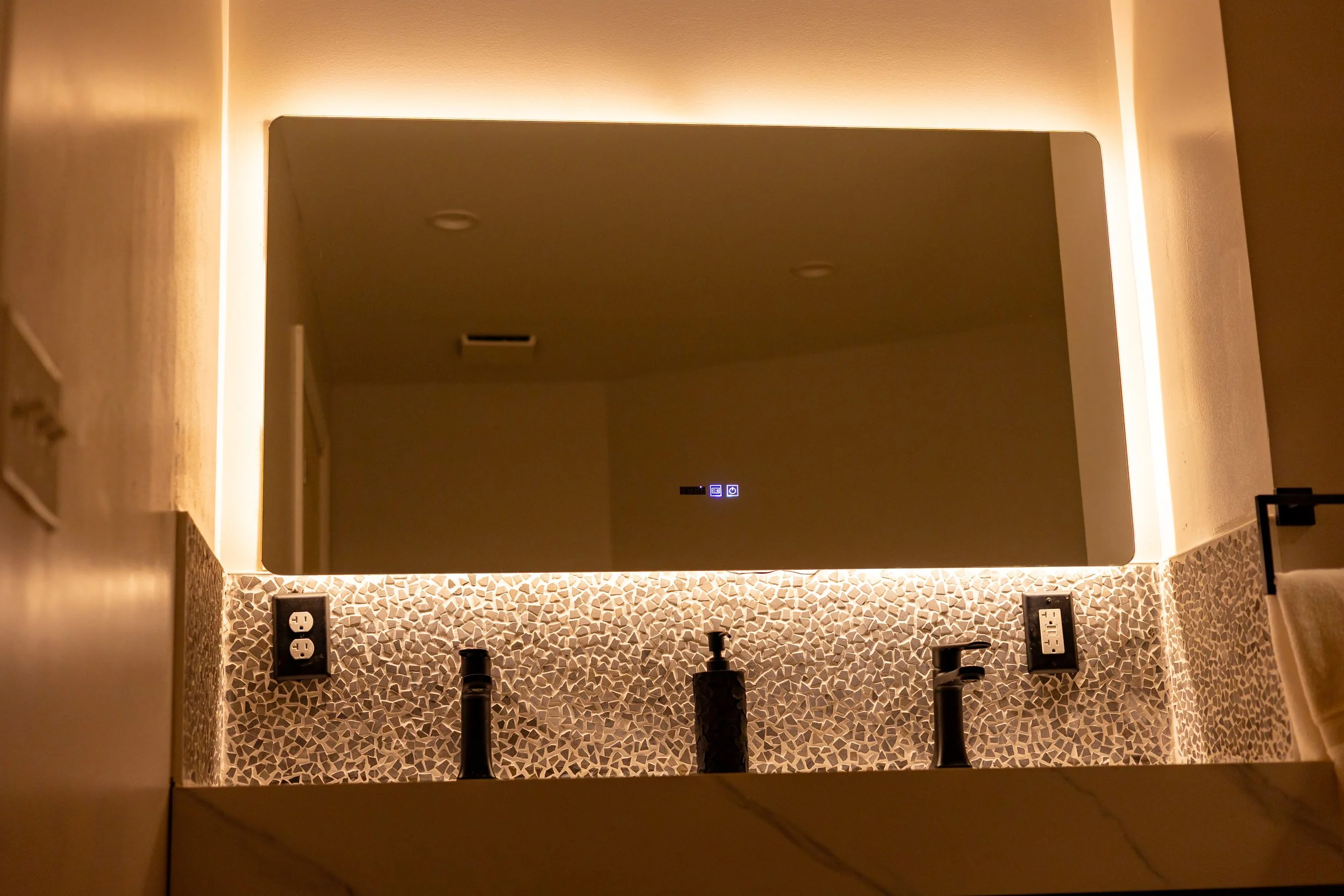 Modern bathroom vanity with mosaic tile countertop, black soap dispenser, faucet, and two electrical outlets, reflected in a large backlit mirror.