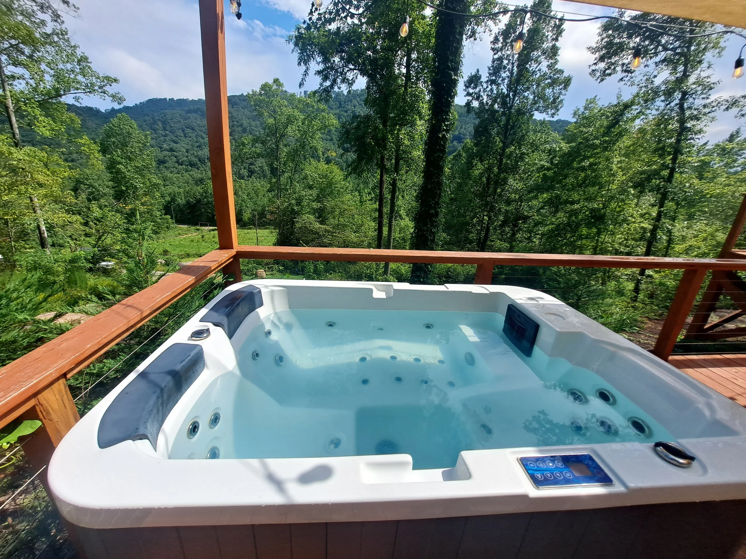 A hot tub on a wooden deck overlooking a lush green mountain landscape with trees and hills, under a partly cloudy sky with string lights hanging above.