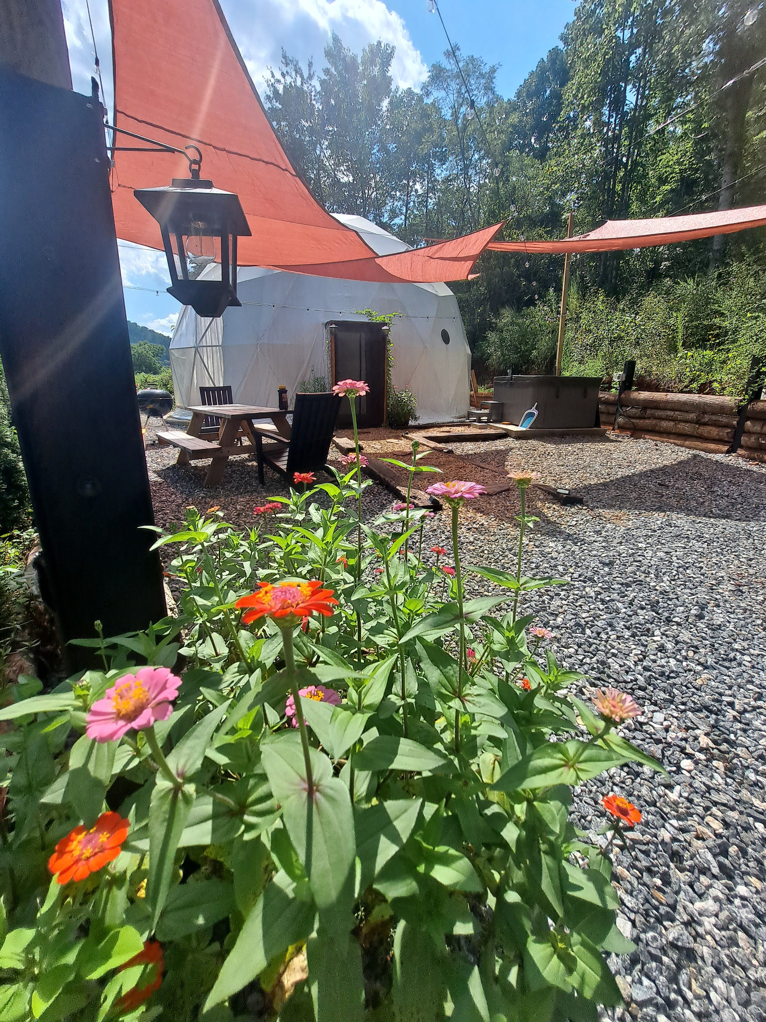 Outdoor seating area with a modern, dome-shaped white yurt, surrounded by flowers, gravel ground, and shaded by outdoor fabric canopies on a sunny day.