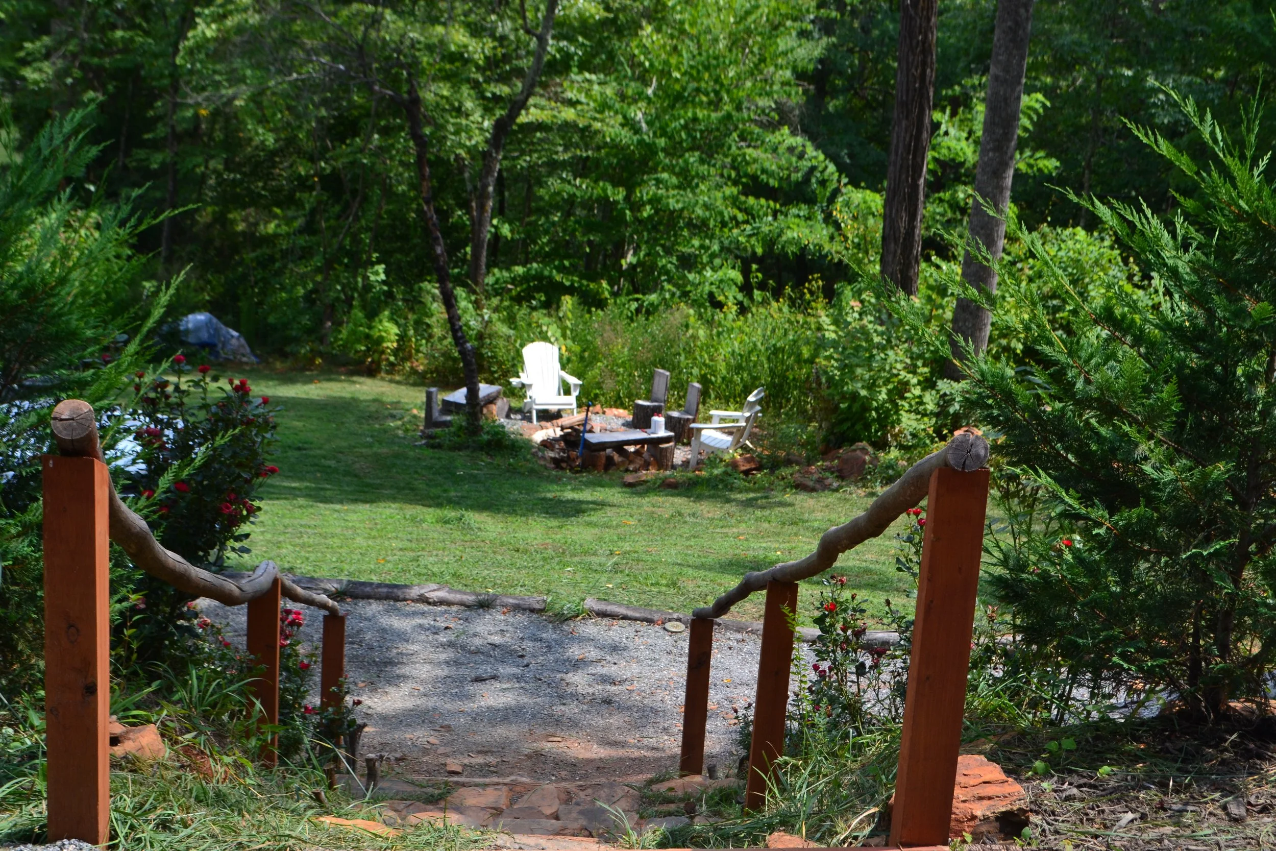 A backyard garden with a staircase leading down to a sitting area with Adirondack chairs around a fire pit, surrounded by lush green trees and bushes.