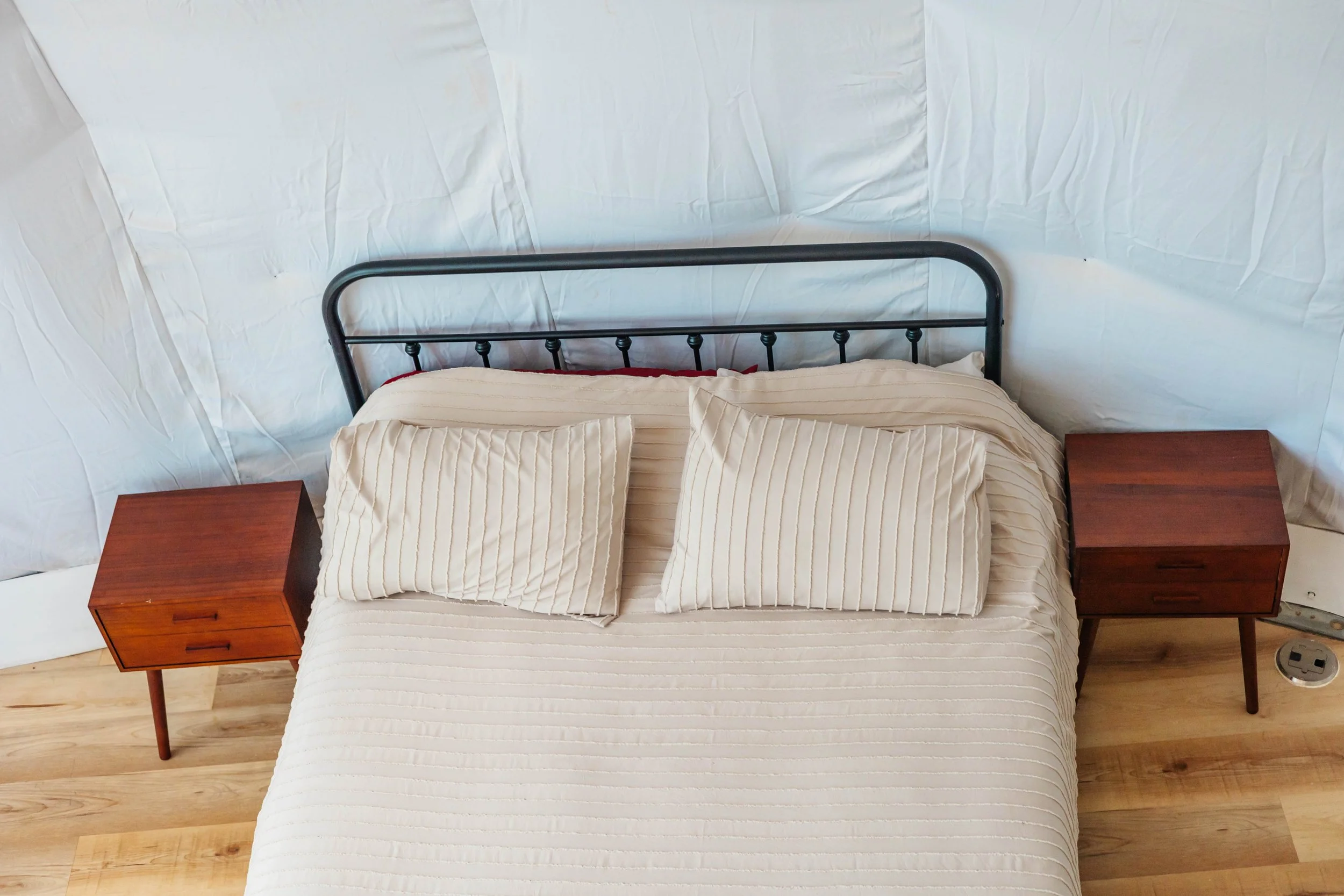Top-down view of a bed with a metal headboard, two beige striped pillows, a matching beige striped bedspread, and wooden nightstands on either side, set against a white background cover.