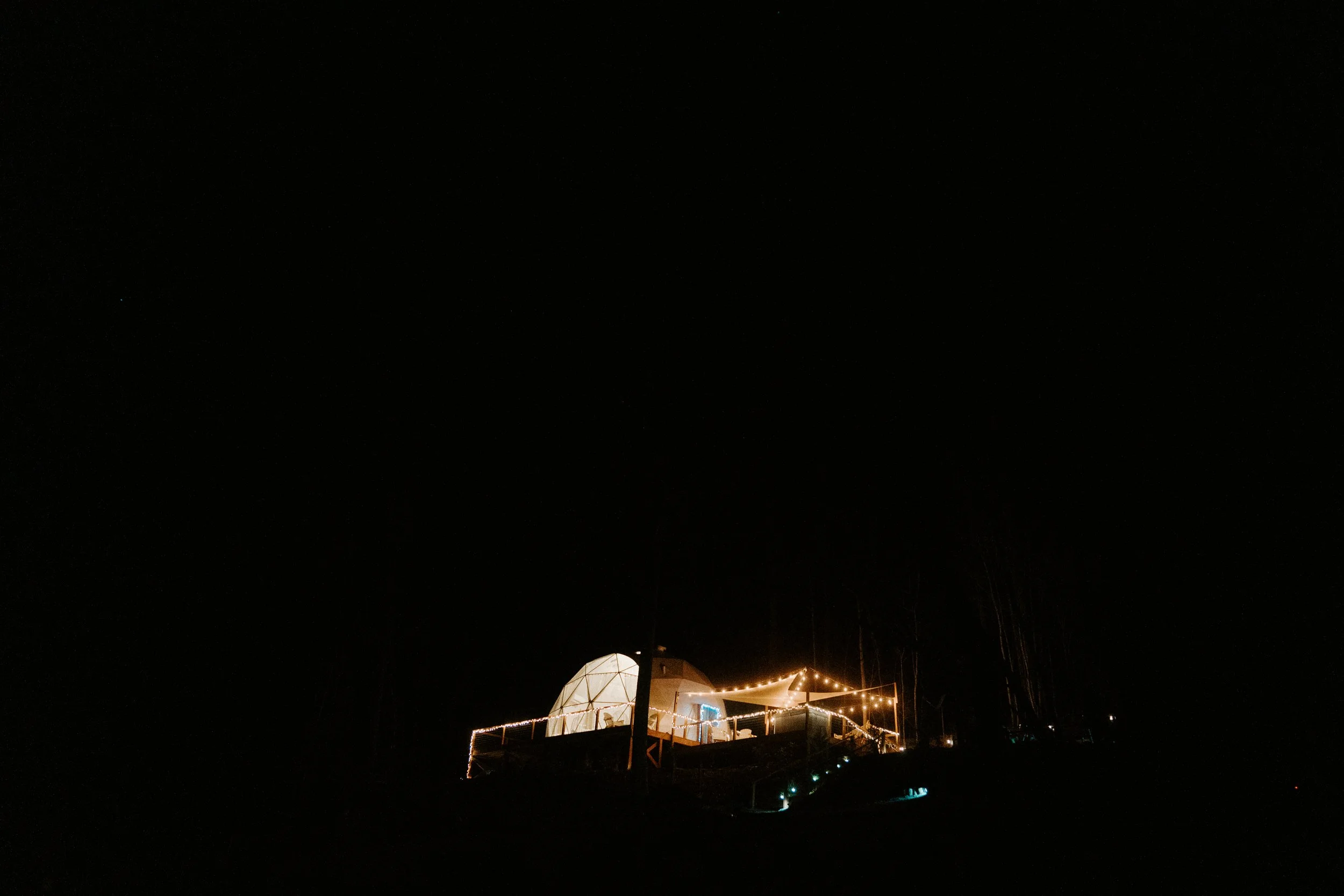 A illuminated dome-shaped tent with string lights on a wooden deck at night, with a dark sky and faint trees in the background.