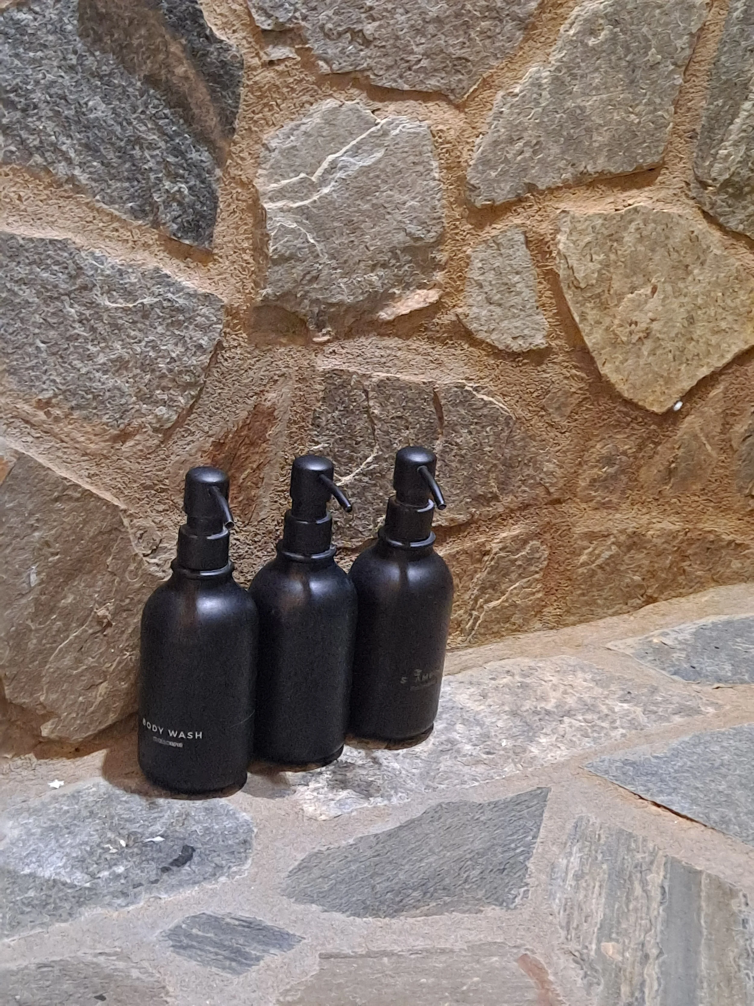 Three black bottles of body wash placed on a stone ledge against a stone wall background.