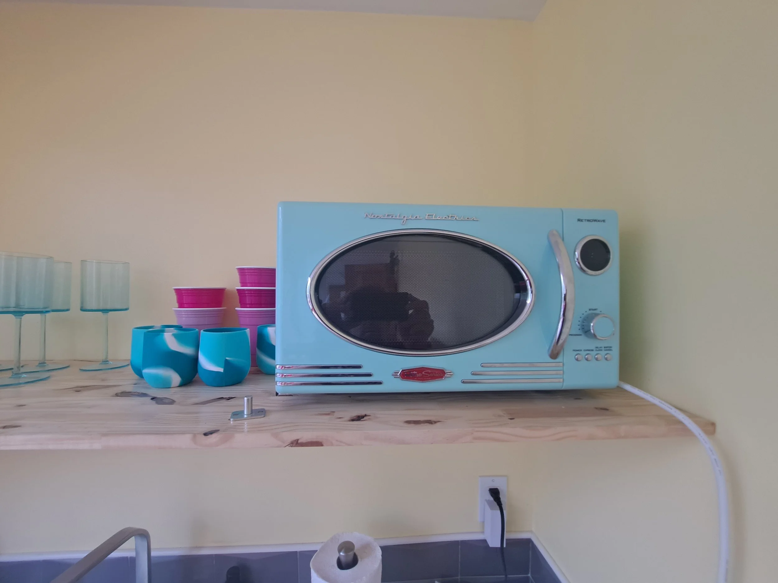 A vintage blue microwave oven with a reflective front, placed on a wooden shelf against a yellow wall, surrounded by colorful cups and glasses.