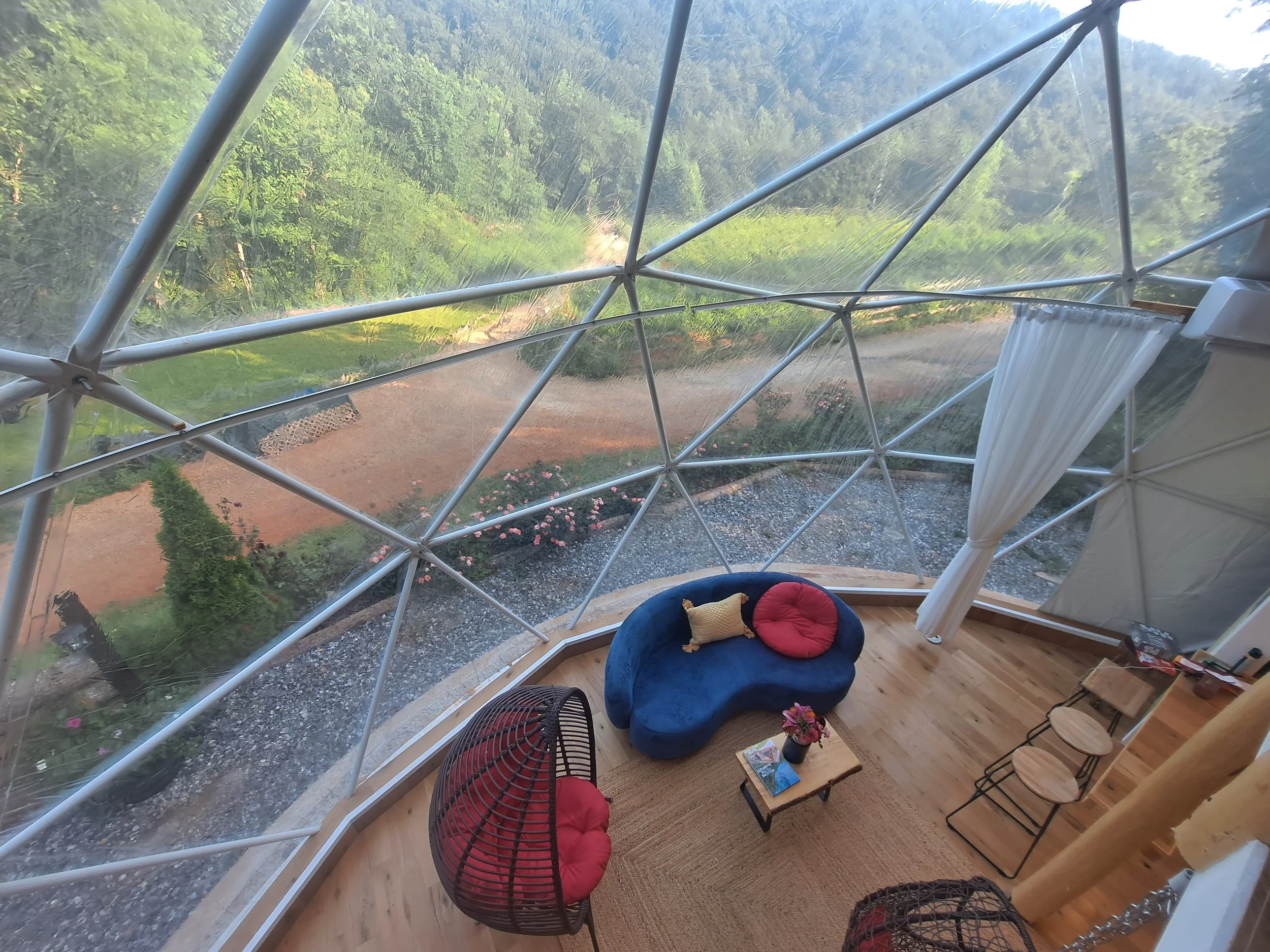 Interior view of a geodesic dome with a curved blue sofa, a red round cushion, a wicker hanging chair with a red cushion, a small wooden table with a flower vase and magazines, and some stacked chairs near a window curtain. The dome has large windows