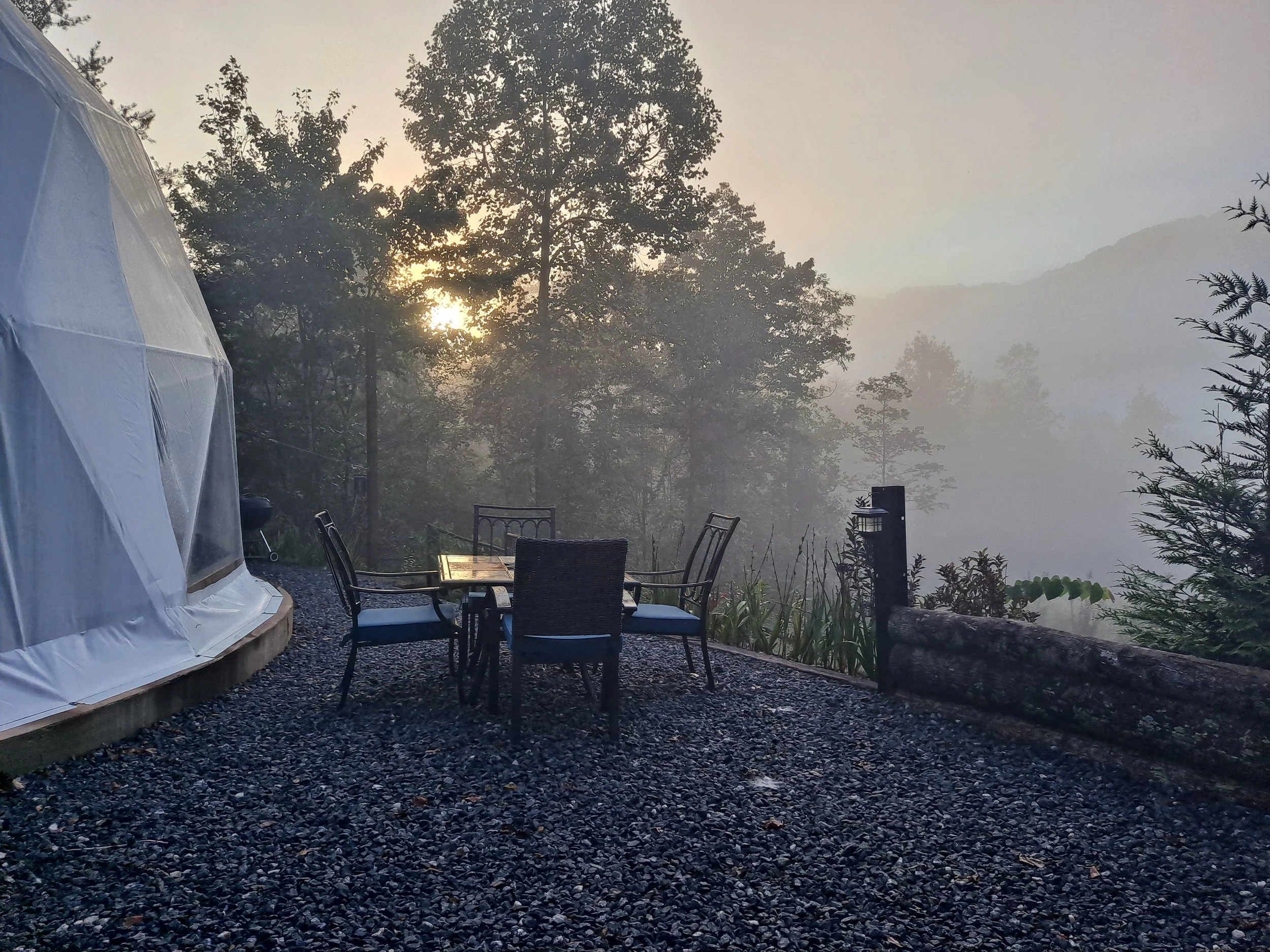 An outdoor gravel patio with a round glass table and four chairs, overlooking a foggy landscape with trees and mountains at sunrise or sunset.