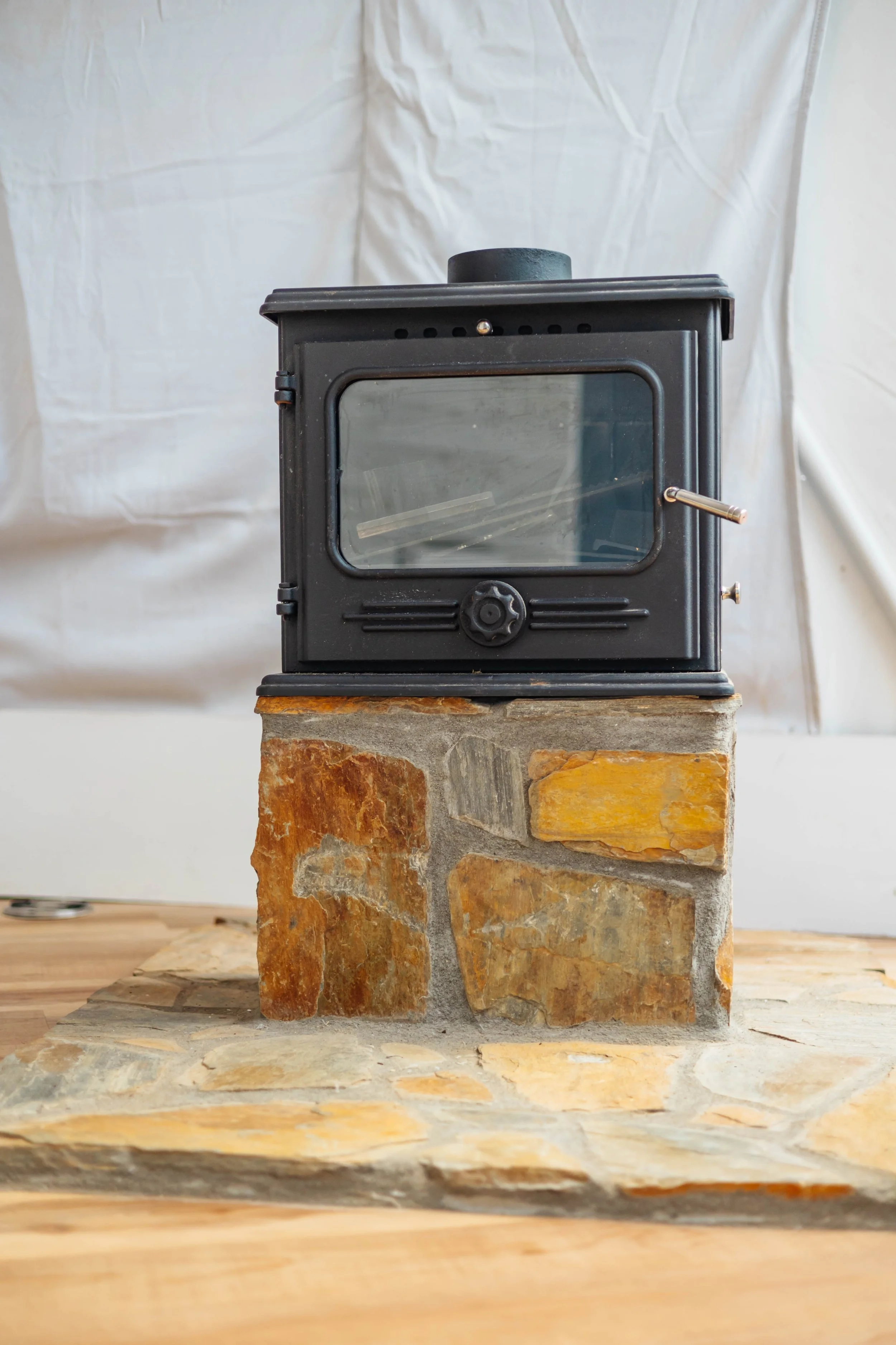 A small black stove with a glass door, sitting on a stone fireplace, on a wooden surface.