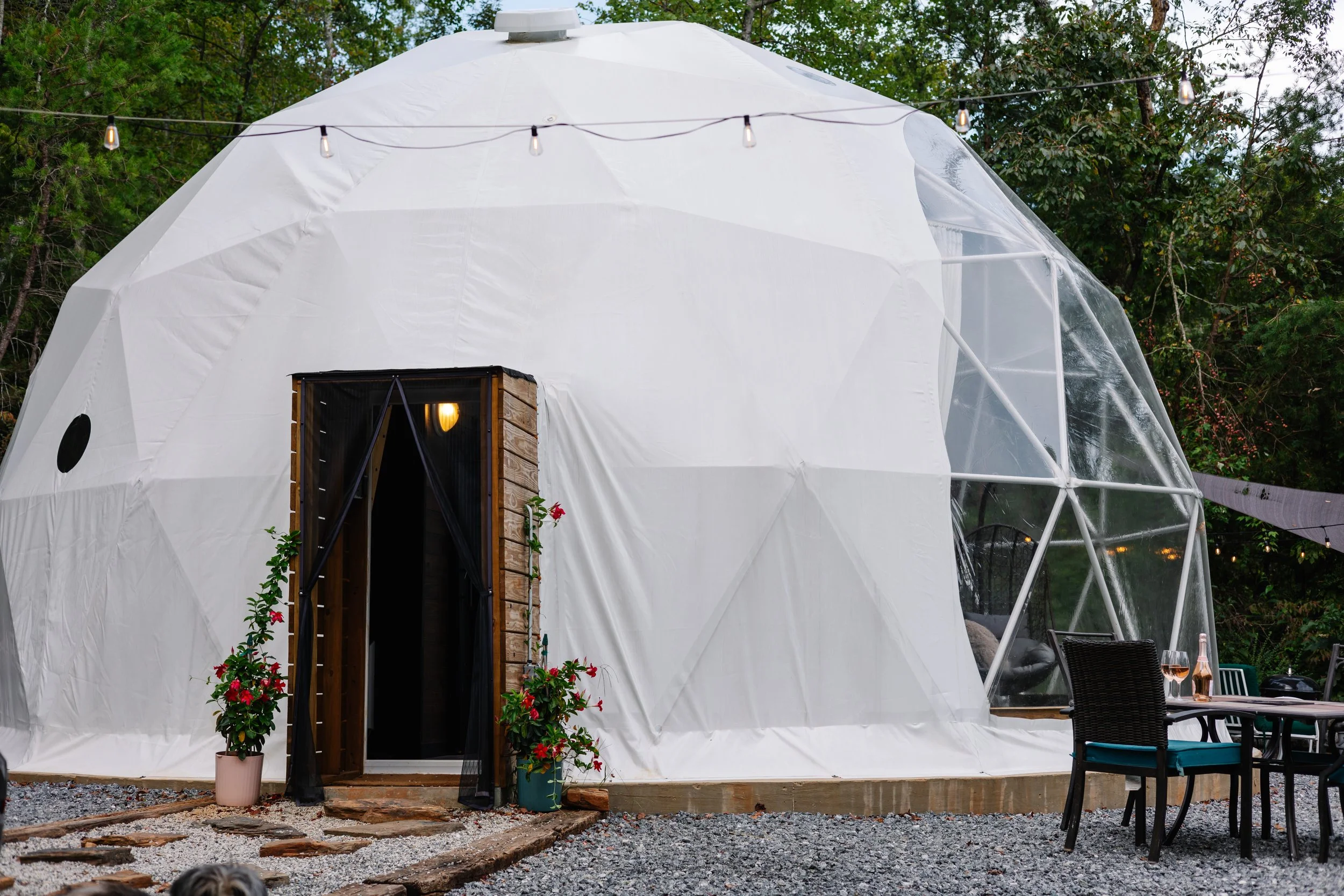 A white geodesic dome structure with transparent sections, decorated with string lights, surrounded by trees and outdoor seating, with potted plants near the entrance.