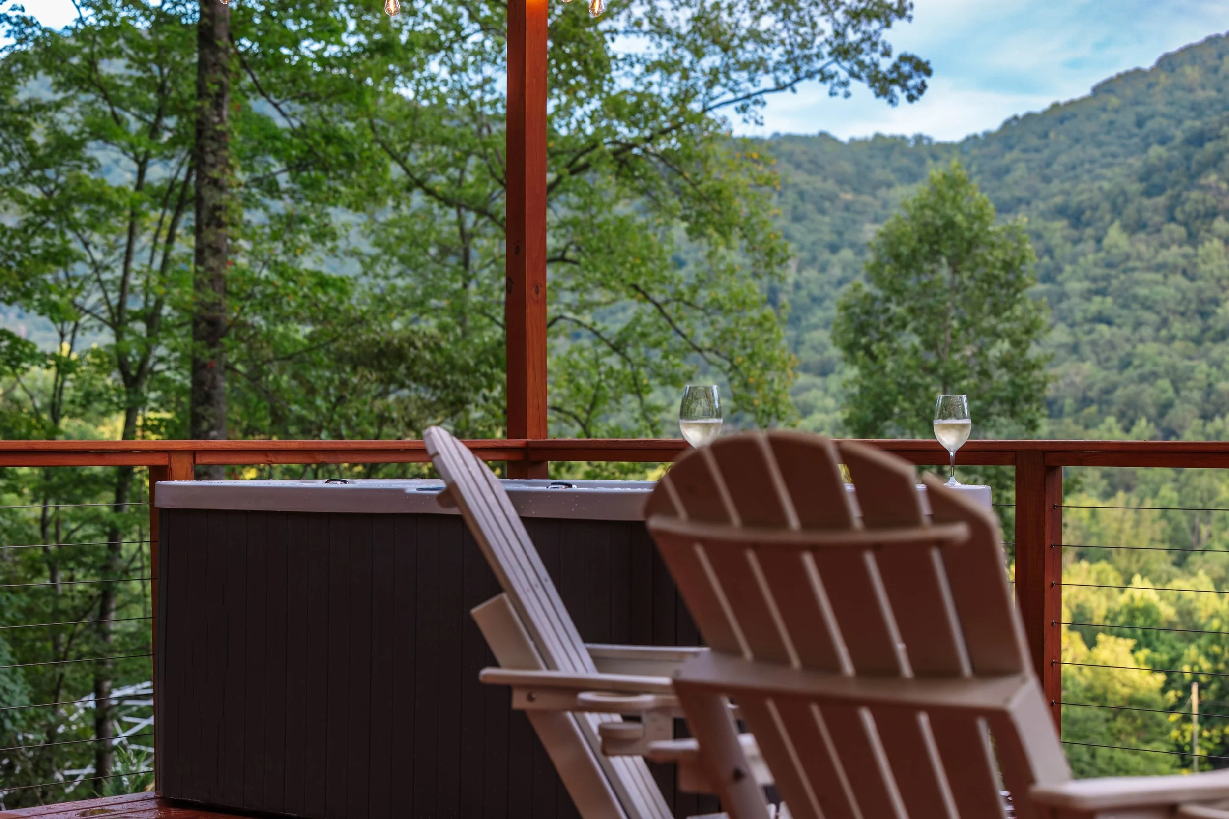 Two glasses of water on a patio railing overlooking a lush green forested mountain landscape with Adirondack chairs in the foreground.