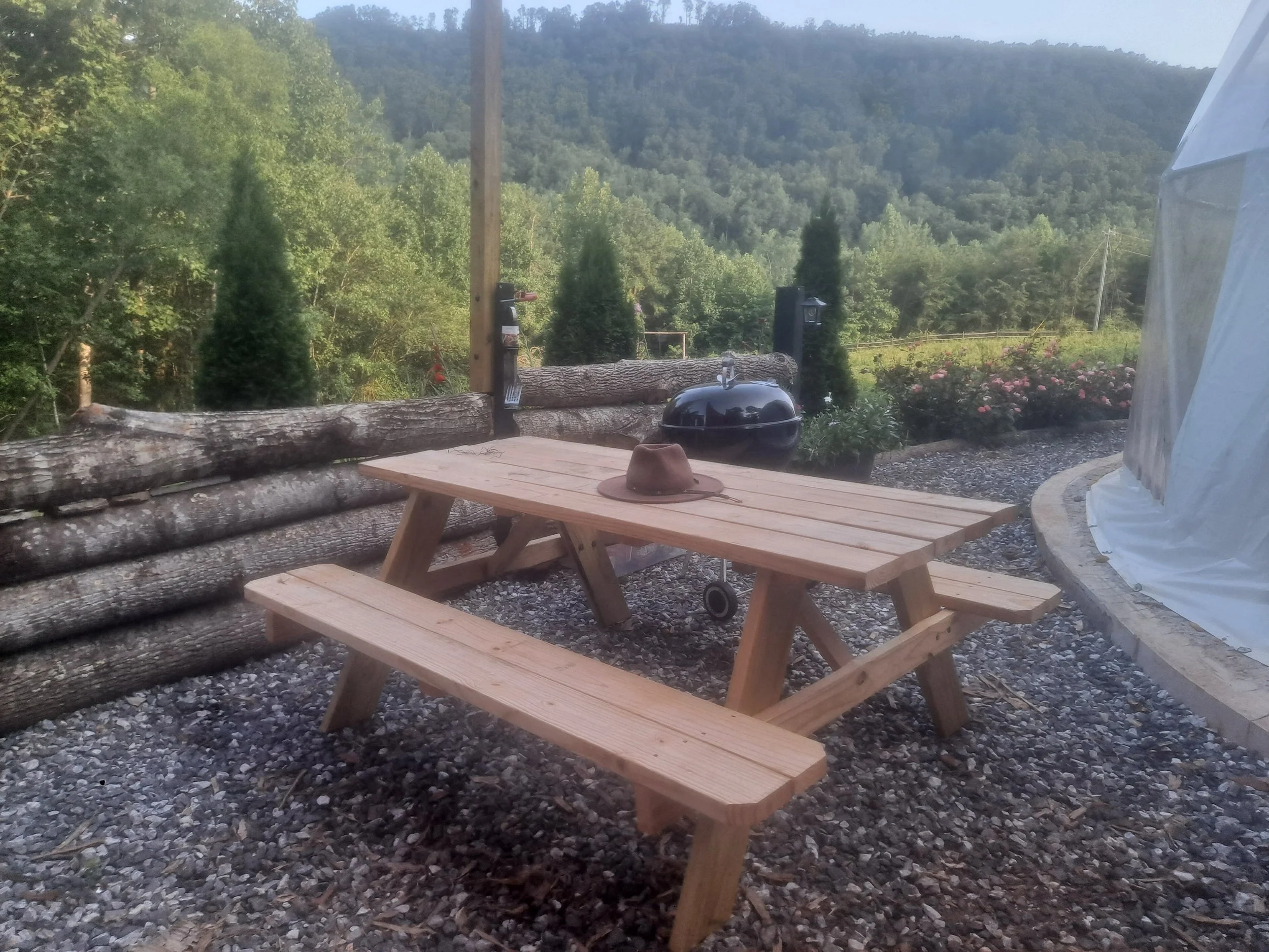 A wooden picnic table with a hat on top, situated outdoors on a gravel surface, with a barbecue grill and lush green trees in the background, during daytime.