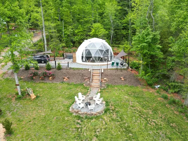 A backyard with a geodesic dome structure, outdoor seating area with chairs and a fire pit, surrounded by green trees and grass.