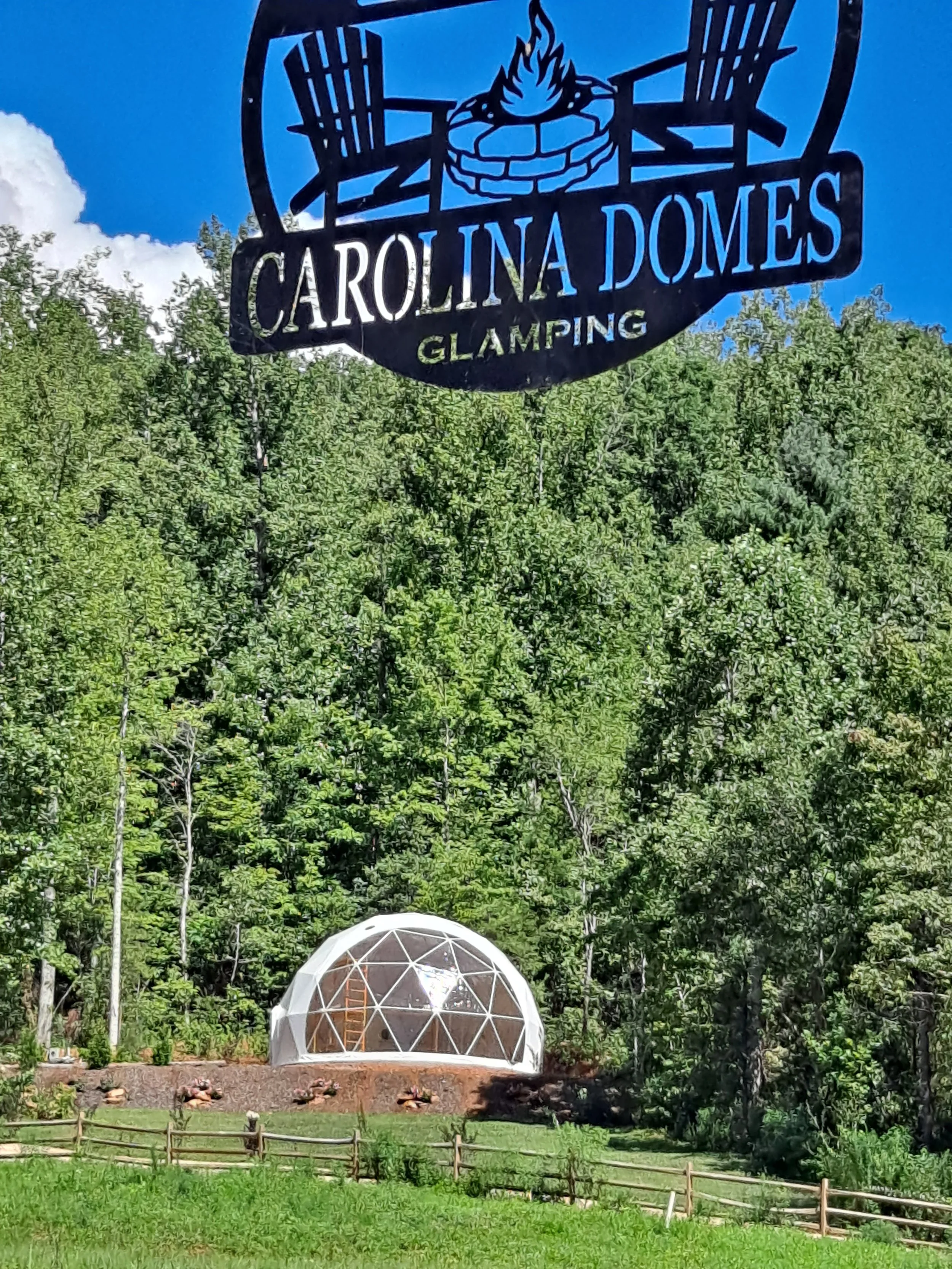Sign for Carolina Domes Glamping above a geodesic dome tent set against a background of green trees and a blue sky.