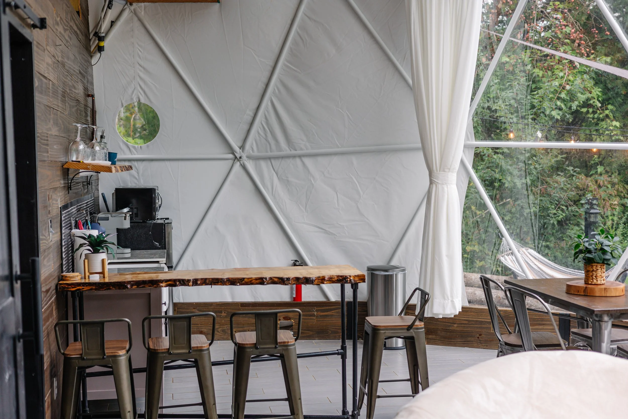 Indoor space with wooden tables and metal chairs, a small kitchen area with a coffee machine, plants, a small round mirror on the wall, and large windows with a view of greenery outside, white curtains partially drawn.