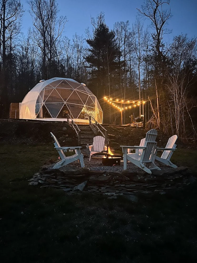 A cozy outdoor scene at dusk with four white Adirondack chairs around a fire pit, a geodesic dome structure with string lights nearby, and a wooded backdrop.
