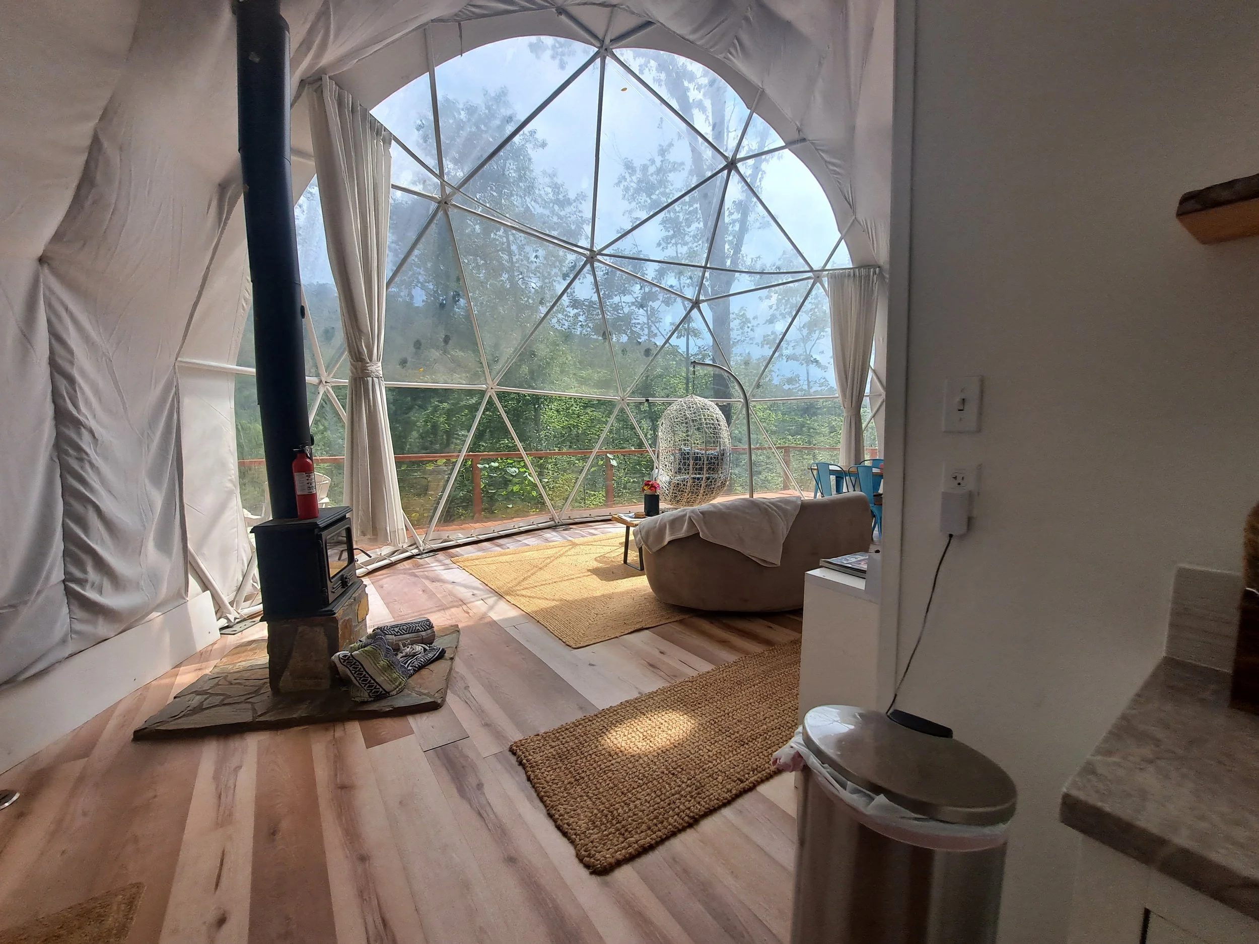 Interior of a dome-shaped living space with large glass windows revealing an outdoor forest scene, a wood stove on a stone platform, a hanging egg chair, a sofa with a blanket, a small table, and a yellow rug.