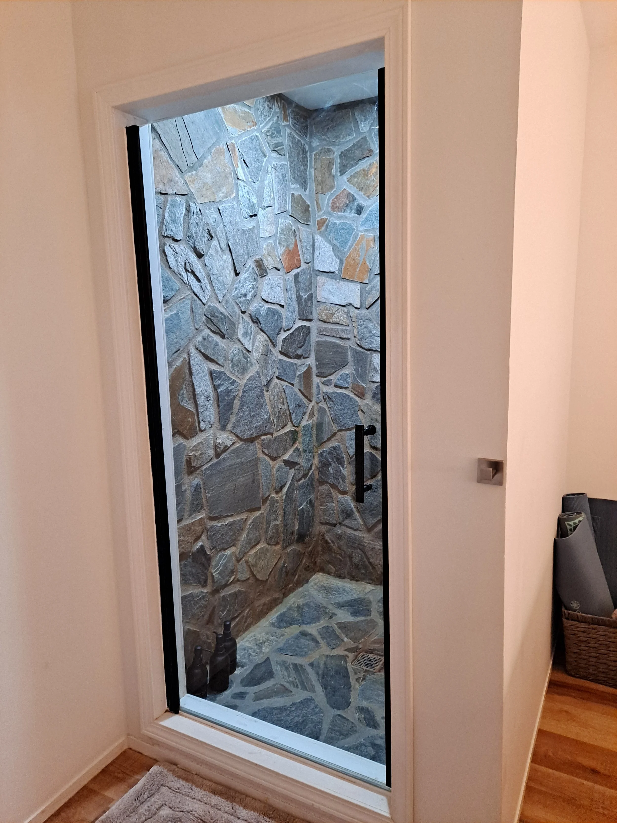 Interior view of a small stone shower with a black door handle, visible from a room with beige walls and wooden floor.