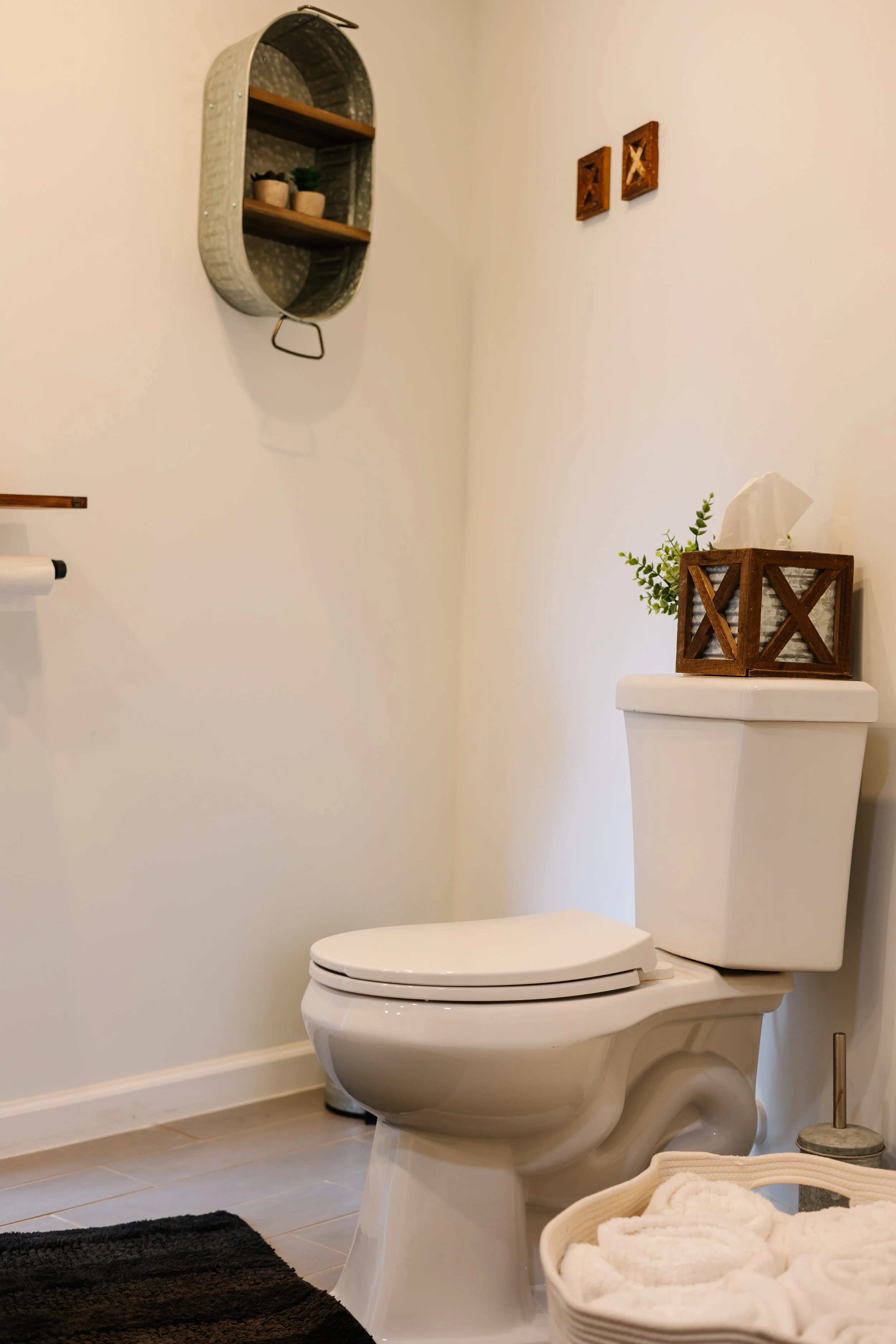 A clean, minimal bathroom with a white toilet, a basket of rolled towels, wall-mounted decorative shelves, and small wall art.