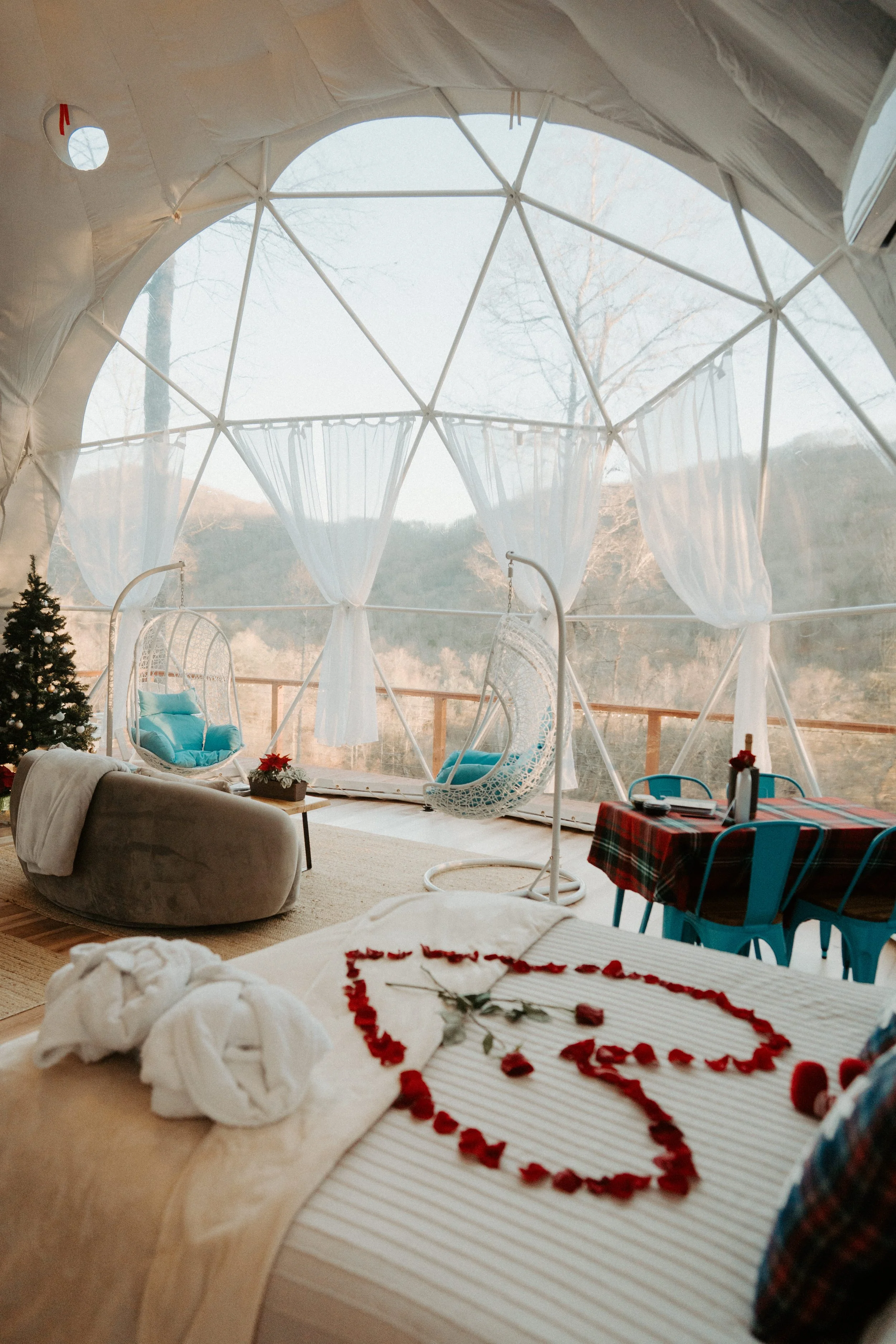 Interior of a cozy domed glamping tent with hanging egg chairs, a small Christmas tree, a couch, a table with a tablecloth, and a bed decorated with rose petals in the shape of a heart and the number 5, overlooking a mountainous landscape.
