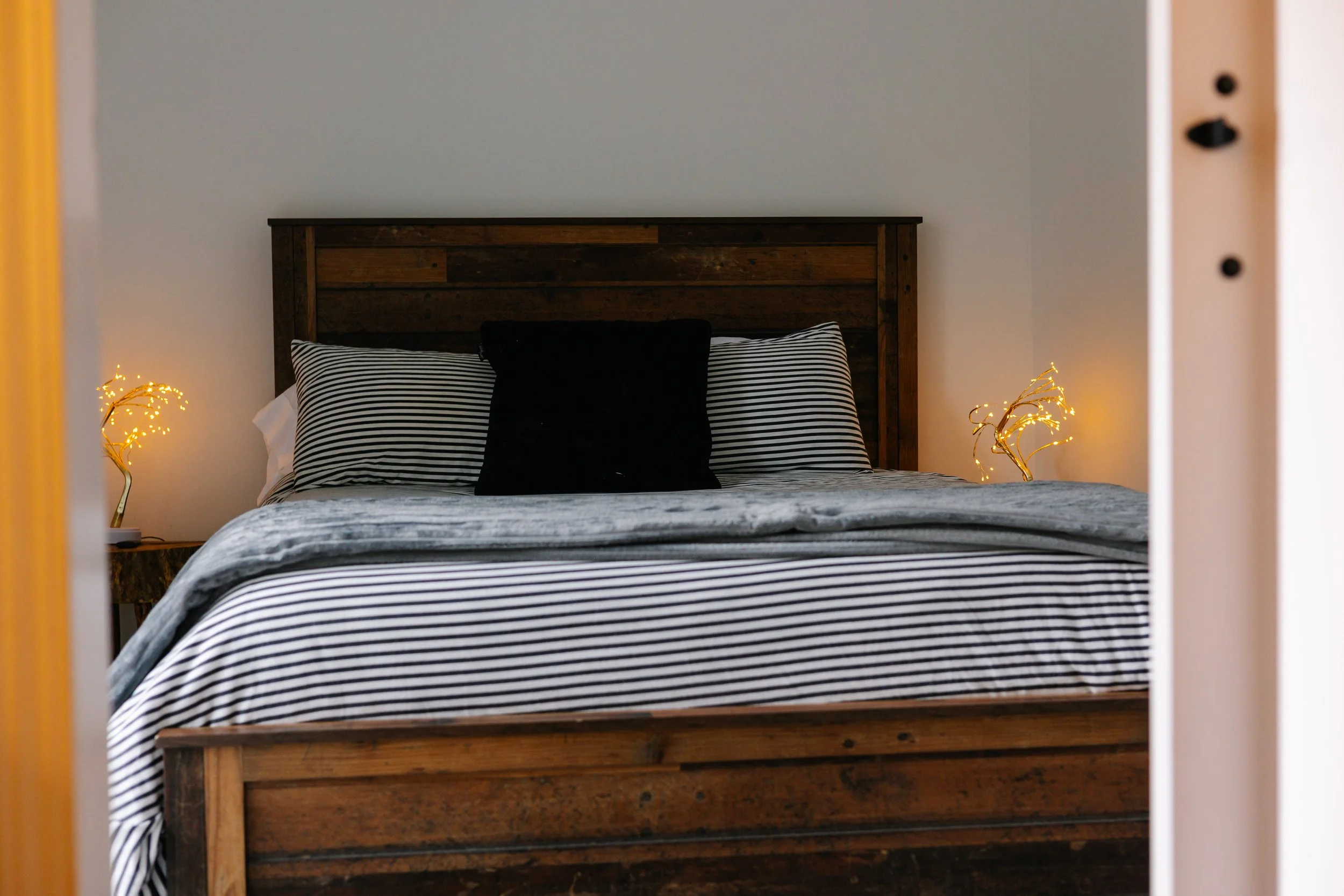 A neatly made bed with striped black and white bedding, a black pillow, and two striped pillows, with warm lighting on bedside lamps shaped like golden branches, viewed through an open door.