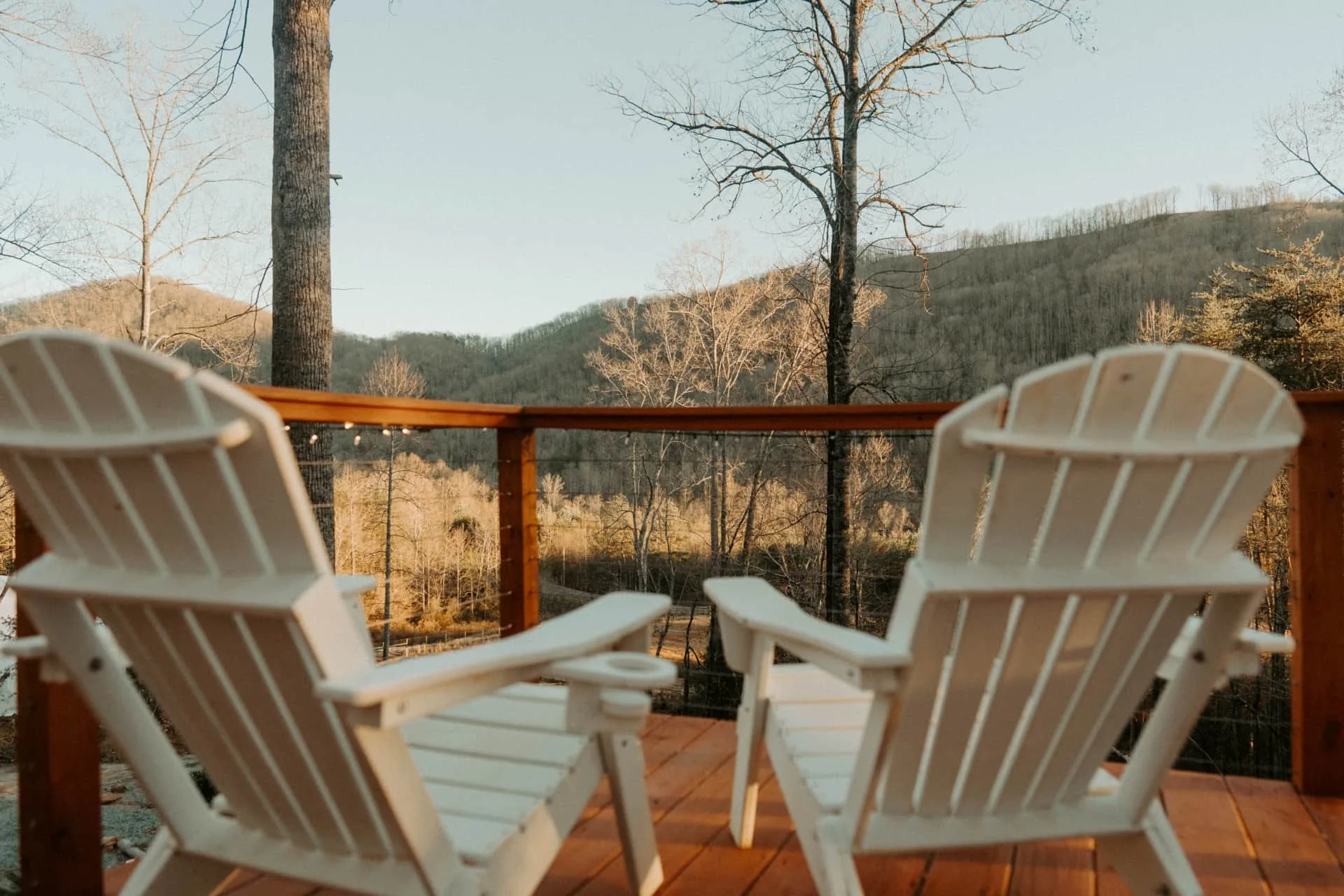 Two white Adirondack chairs on a wooden deck overlook a wooded landscape with mountains in the distance during late afternoon.