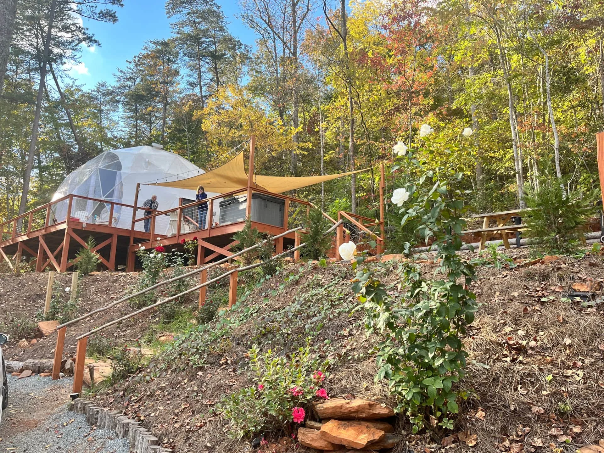 A geodesic dome with a deck is situated in a wooded area with trees and colorful autumn leaves, featuring two people standing on the deck near a shaded area with string lights.