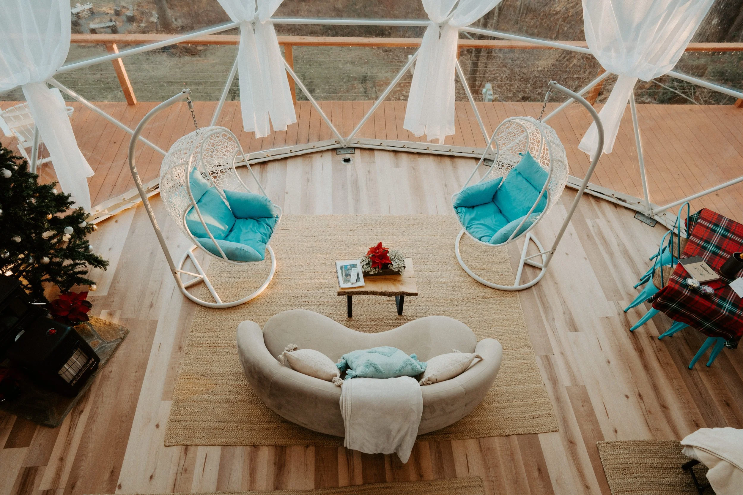 A cozy living area with a beige curved sofa with pillows, a small wooden coffee table with a poinsettia plant, two hanging patio chairs with blue cushions, and a dining table with a tartan tablecloth. There is a Christmas tree with ornaments in the c