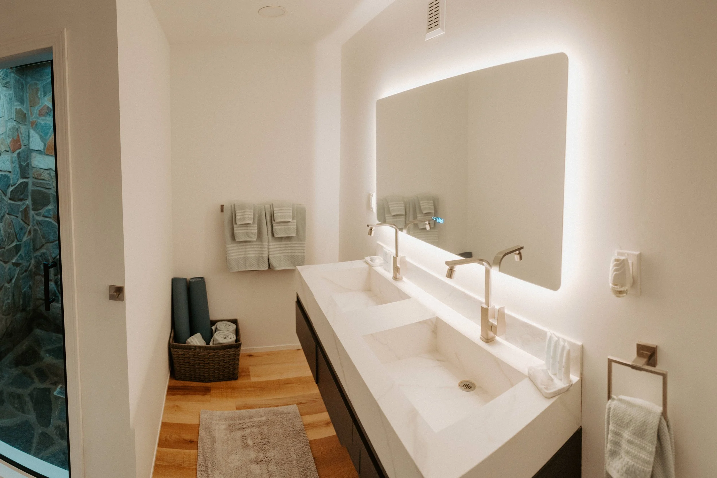 A modern bathroom with a double white sink vanity, large backlit mirror, light-colored towels, and a wooden floor. There is a basket with rolled towels and a shower with a stone interior visible on the left.