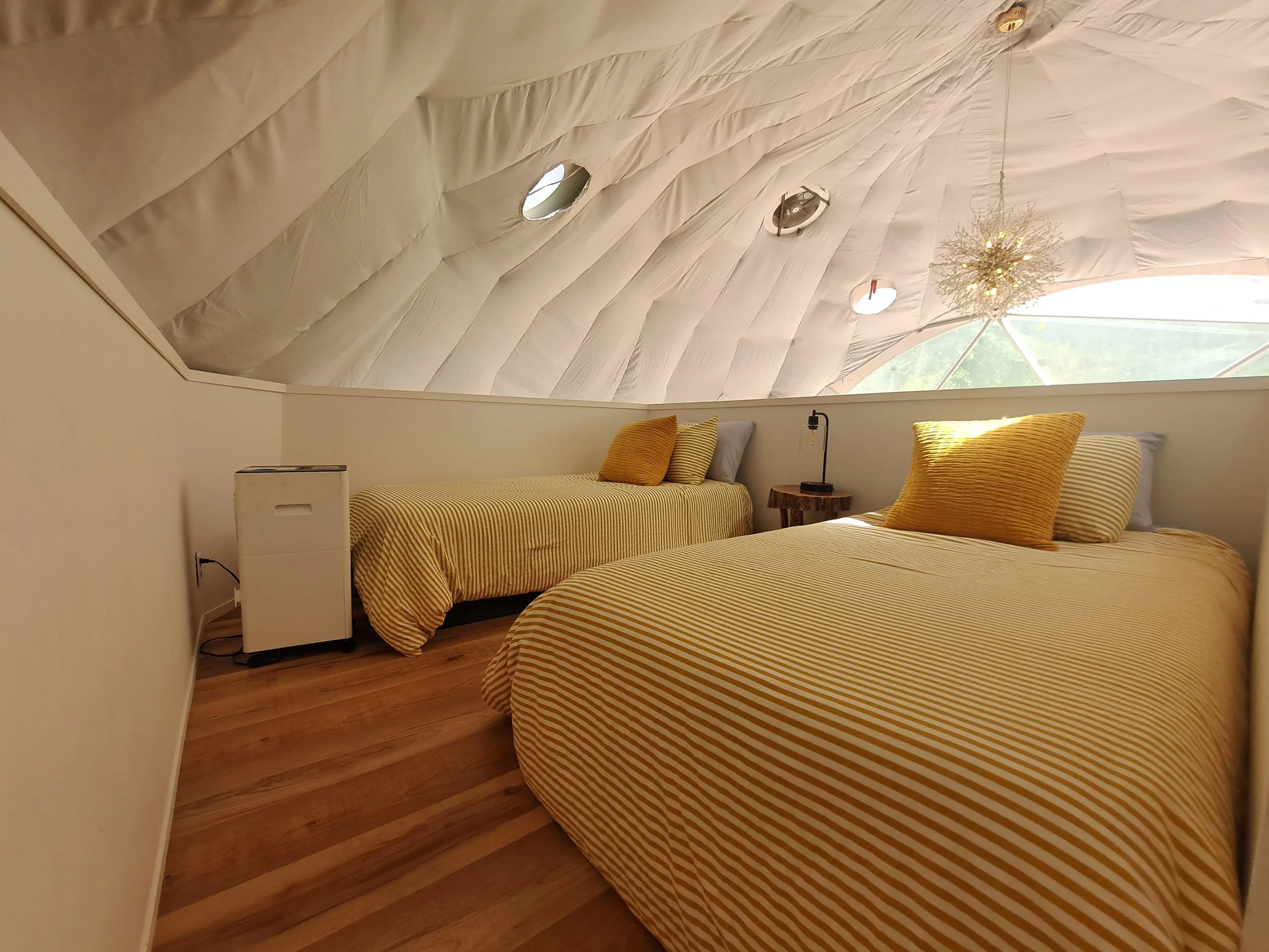 A cozy attic bedroom with two twin beds covered in yellow and white striped bedding, a small wooden nightstand, a black desk lamp, a decorative light fixture hanging from the ceiling, and a large angled window with a view of trees outside.