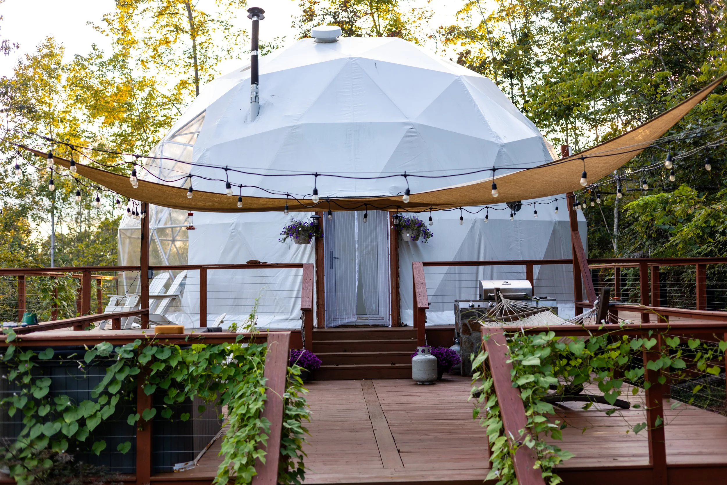 A modern white geodesic dome on a wooden deck surrounded by trees, with string lights and outdoor furniture.