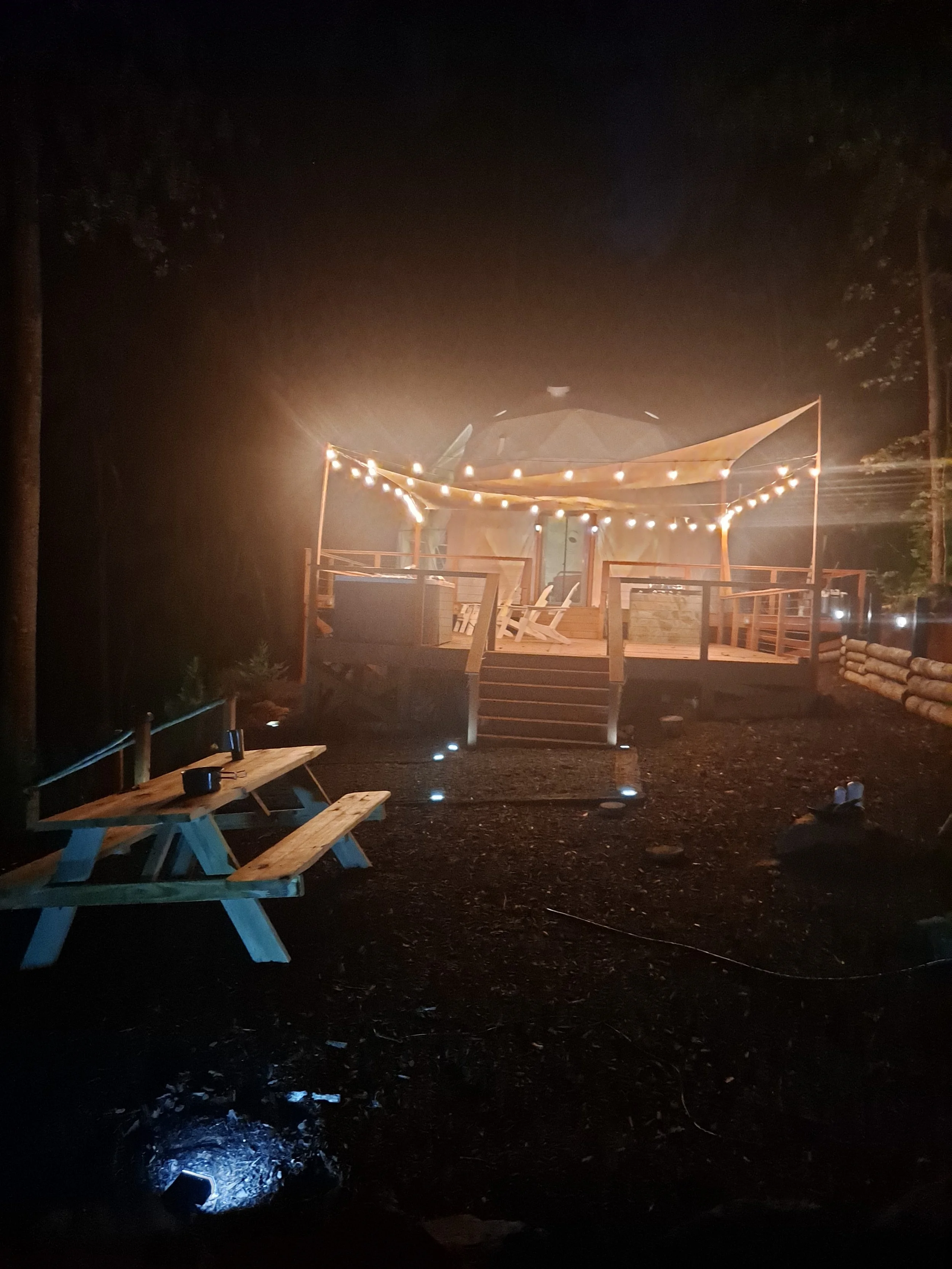 Nighttime outdoor scene with a deck and tent illuminated by string lights. There are Adirondack chairs on the deck and a picnic table nearby.