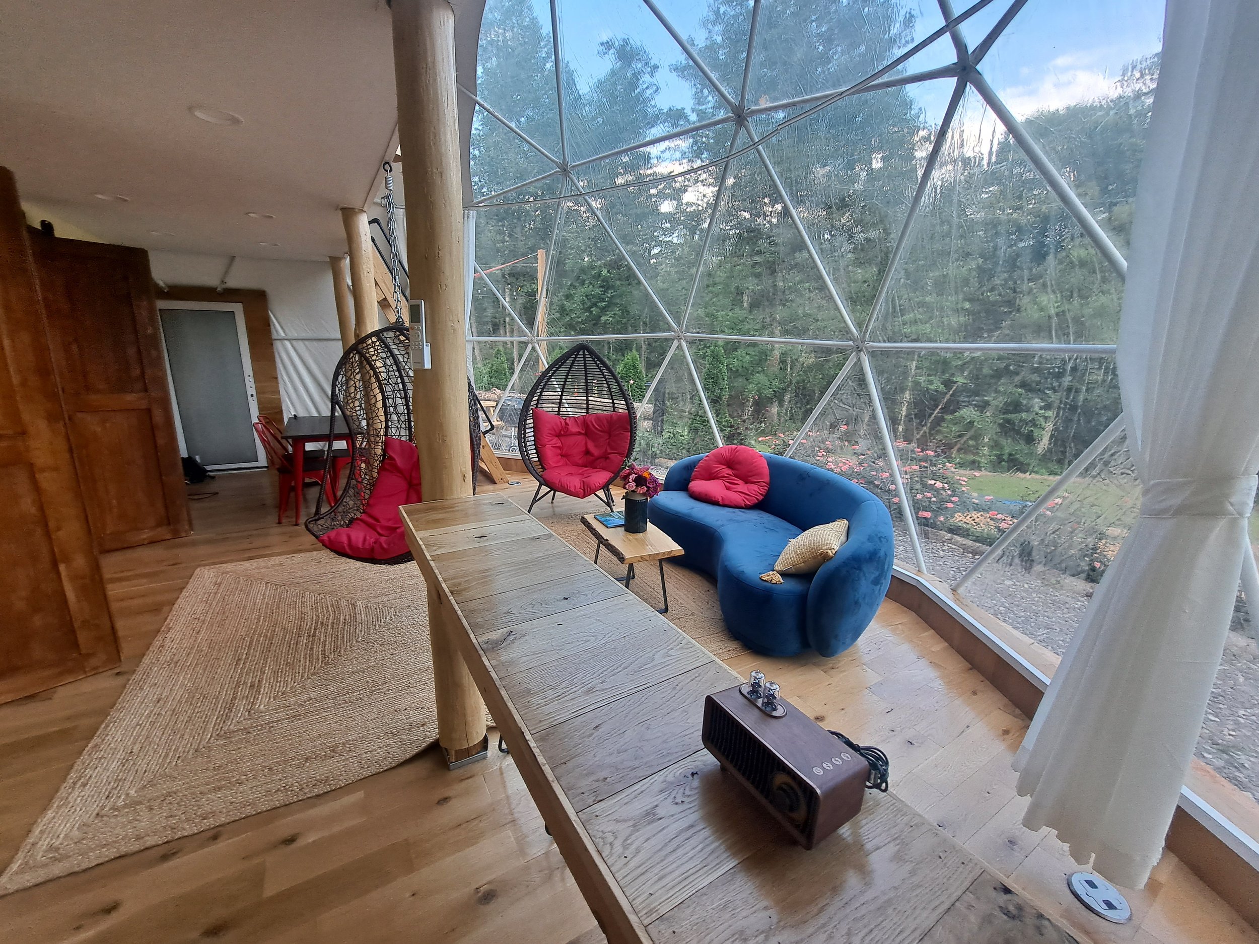 A cozy indoor space with large geodesic glass windows showing a garden outside. The room has a wooden floor, a beige rug, and a wooden table. There are two red hanging egg chairs with cushions, a blue sofa with pillows, and a small side table with a 