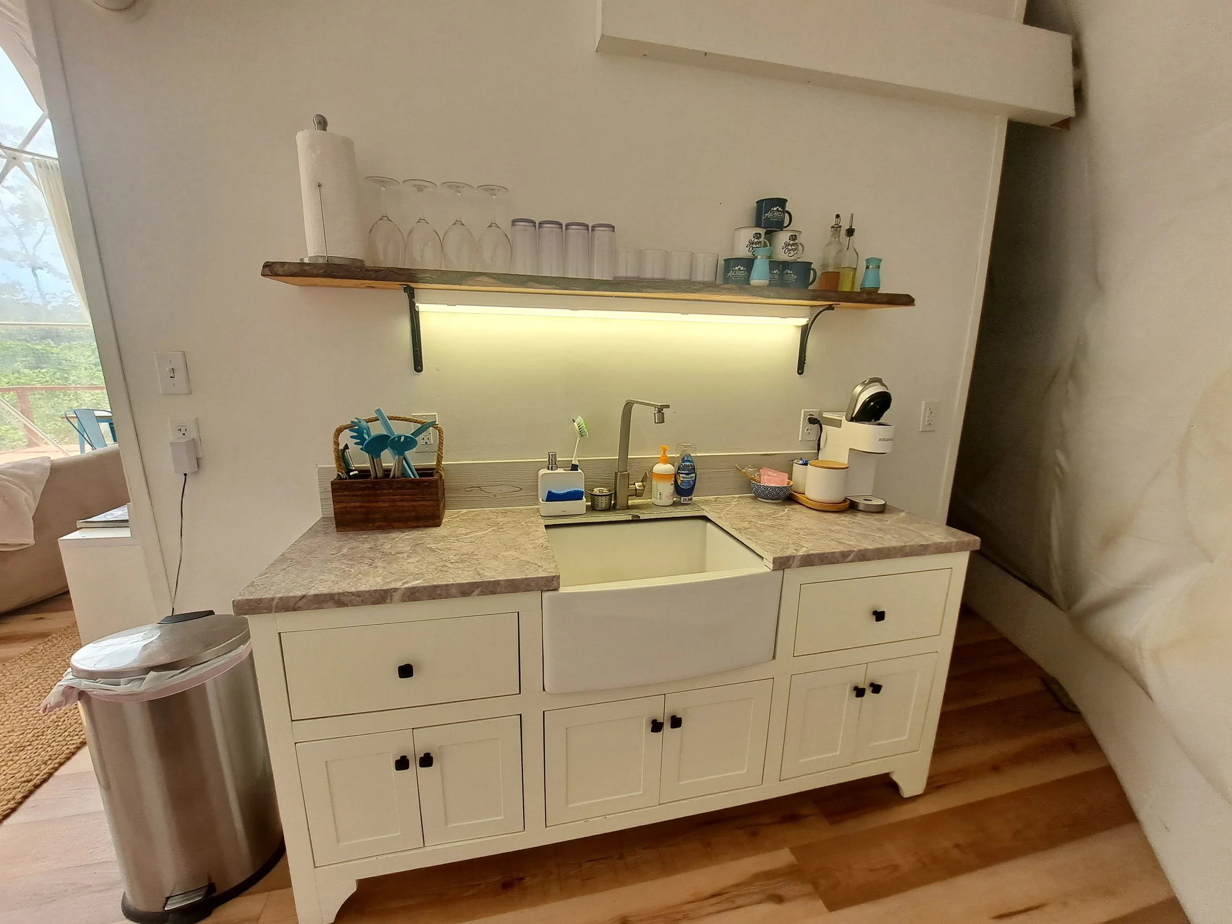 Kitchen sink area with a beige cabinet, a marble countertop, a wooden shelf with glasses, cups, and bottles, a basket of kitchen utensils, and various small items.