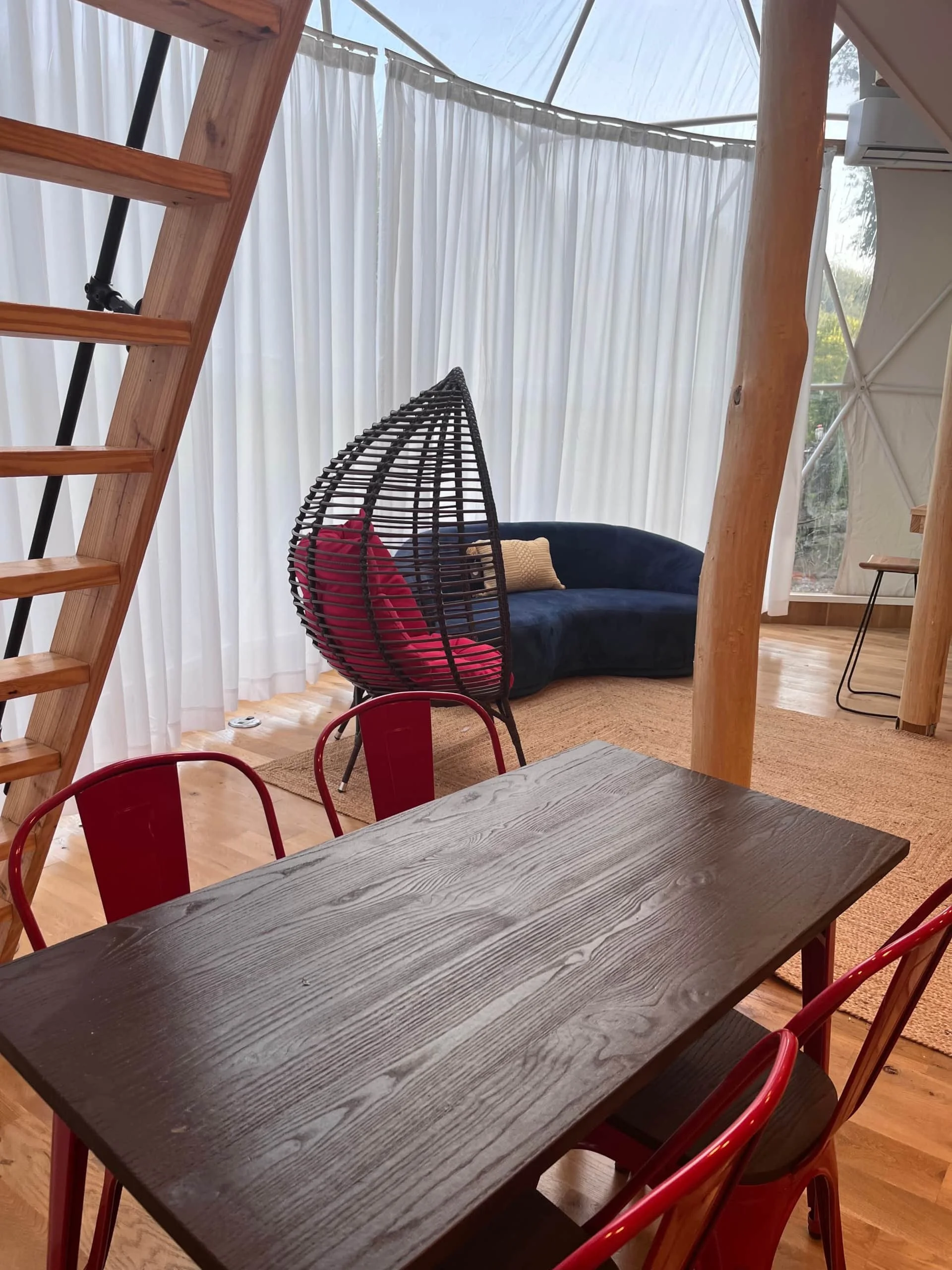 Interior of a cozy room with a wooden table and red chairs, a black wicker hanging chair with pillows, a navy sofa, white curtains, and a visible outdoor scene through a window.