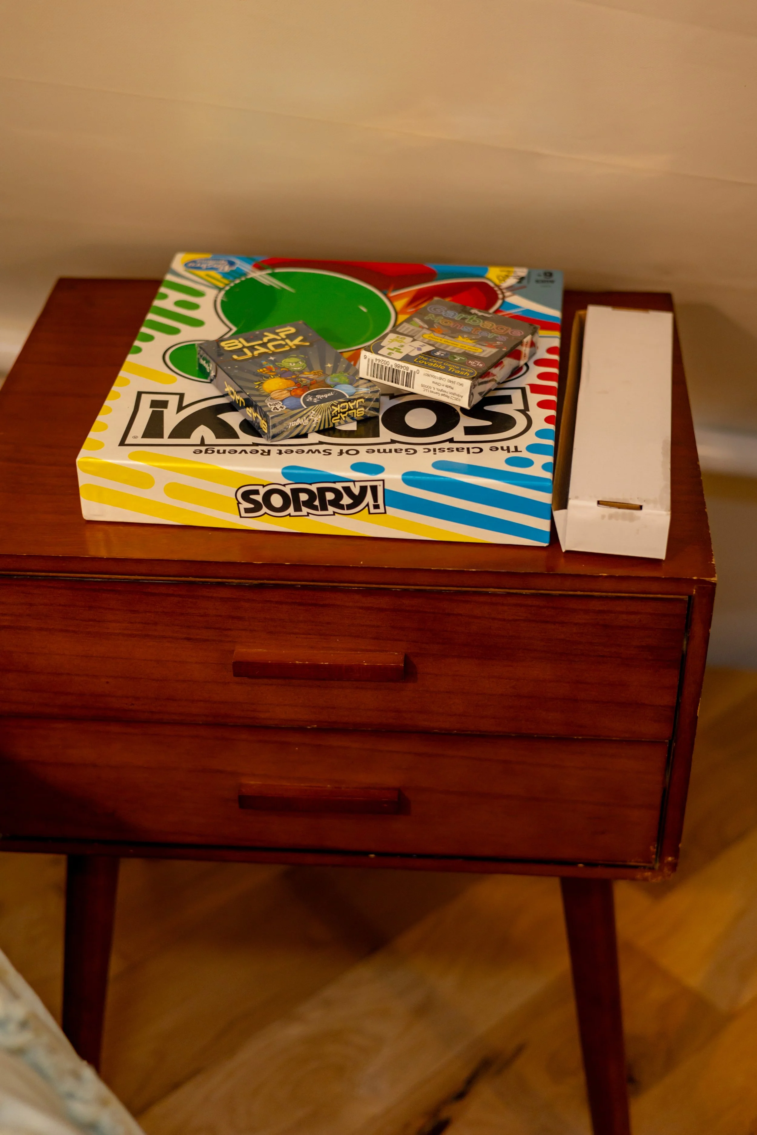 A wooden nightstand with two drawers, on top of which there is a Sorry! board game, a box of vintage Top Trumps cards, and a white rectangular box.