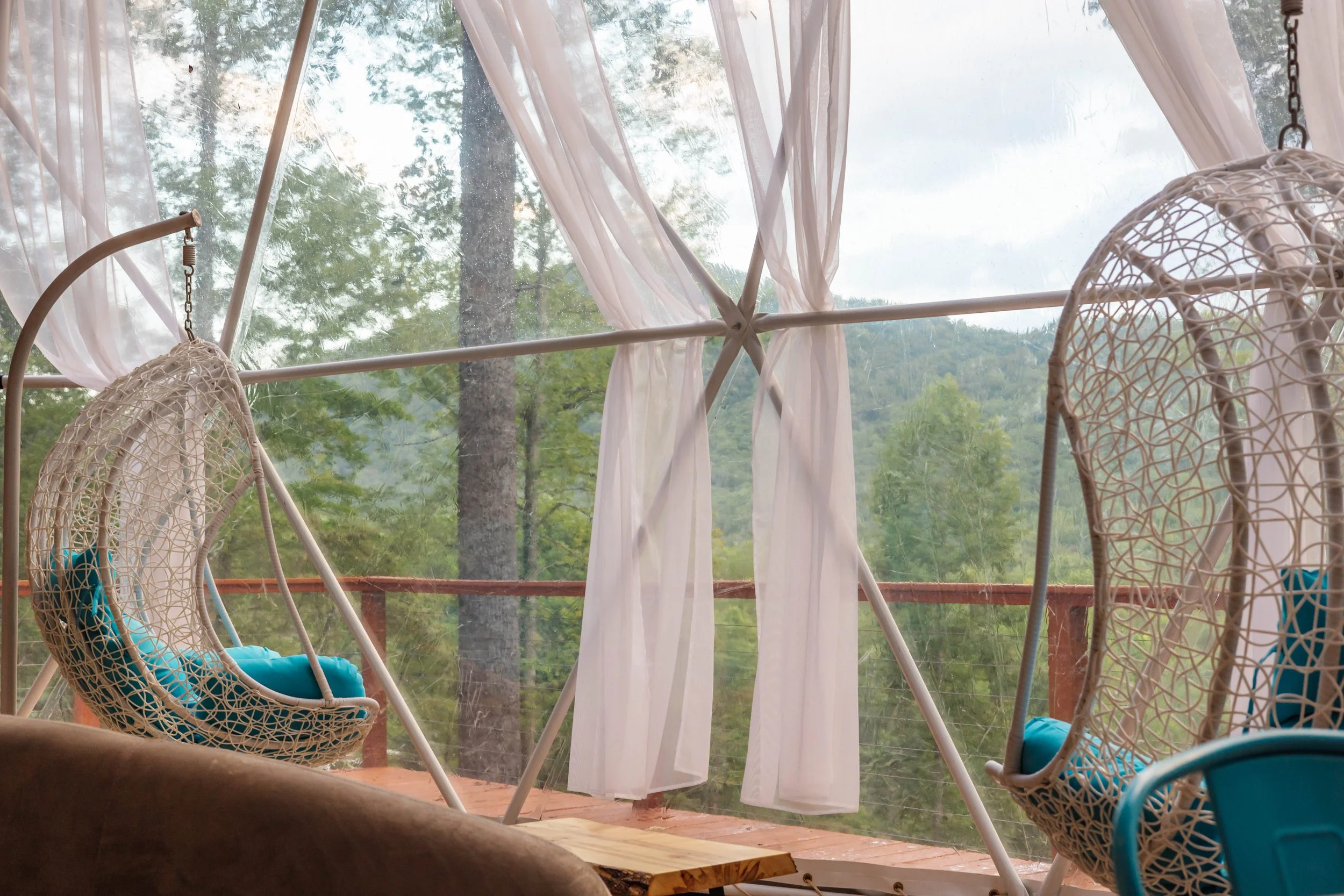 A cozy outdoor porch with two hanging egg chairs with blue cushions, white curtains, and a view of green trees and mountains in the background.