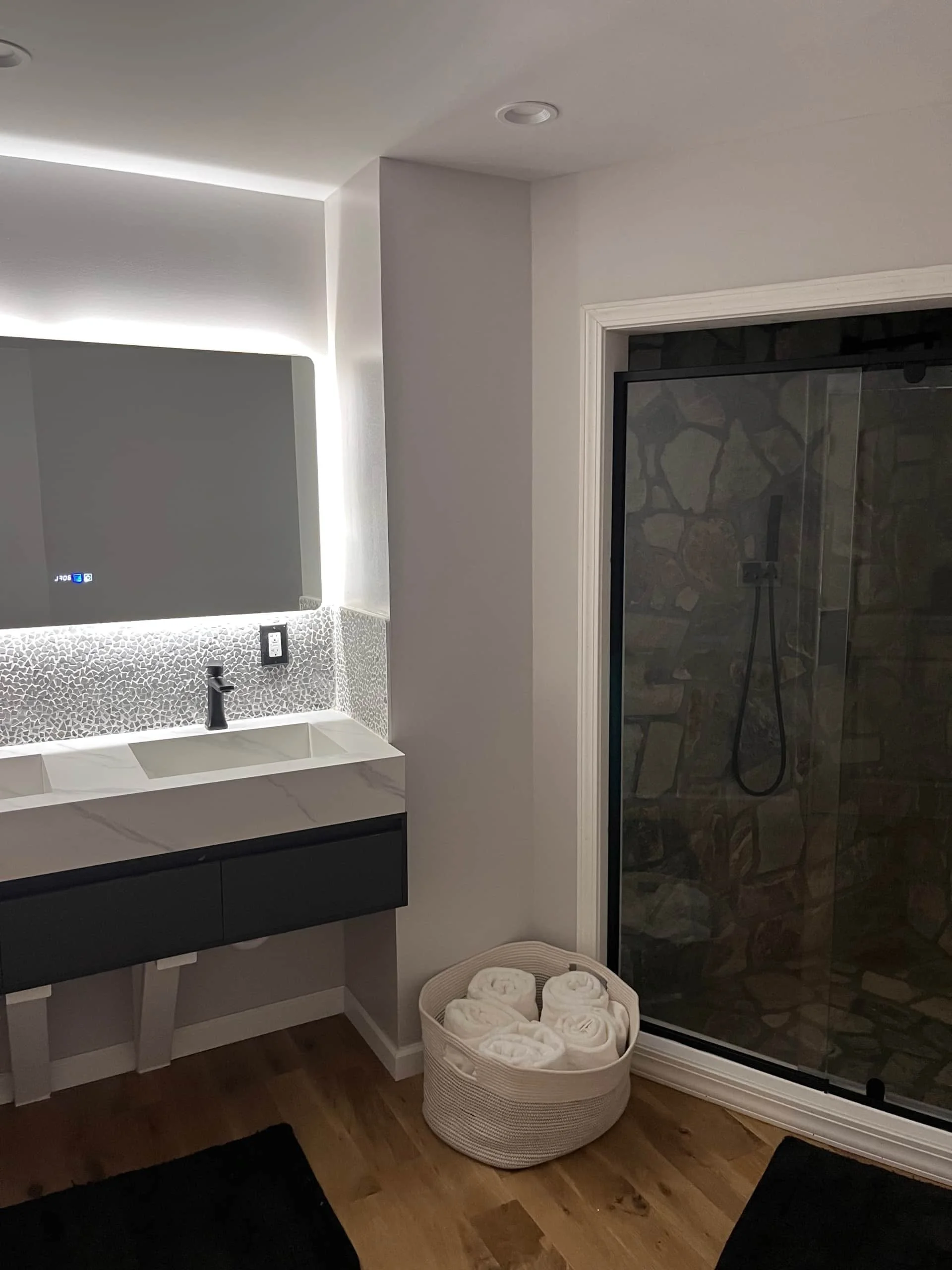 Bathroom with a modern sink and mirror, a basket of neatly rolled white towels, and a glass shower enclosure with a stone interior.