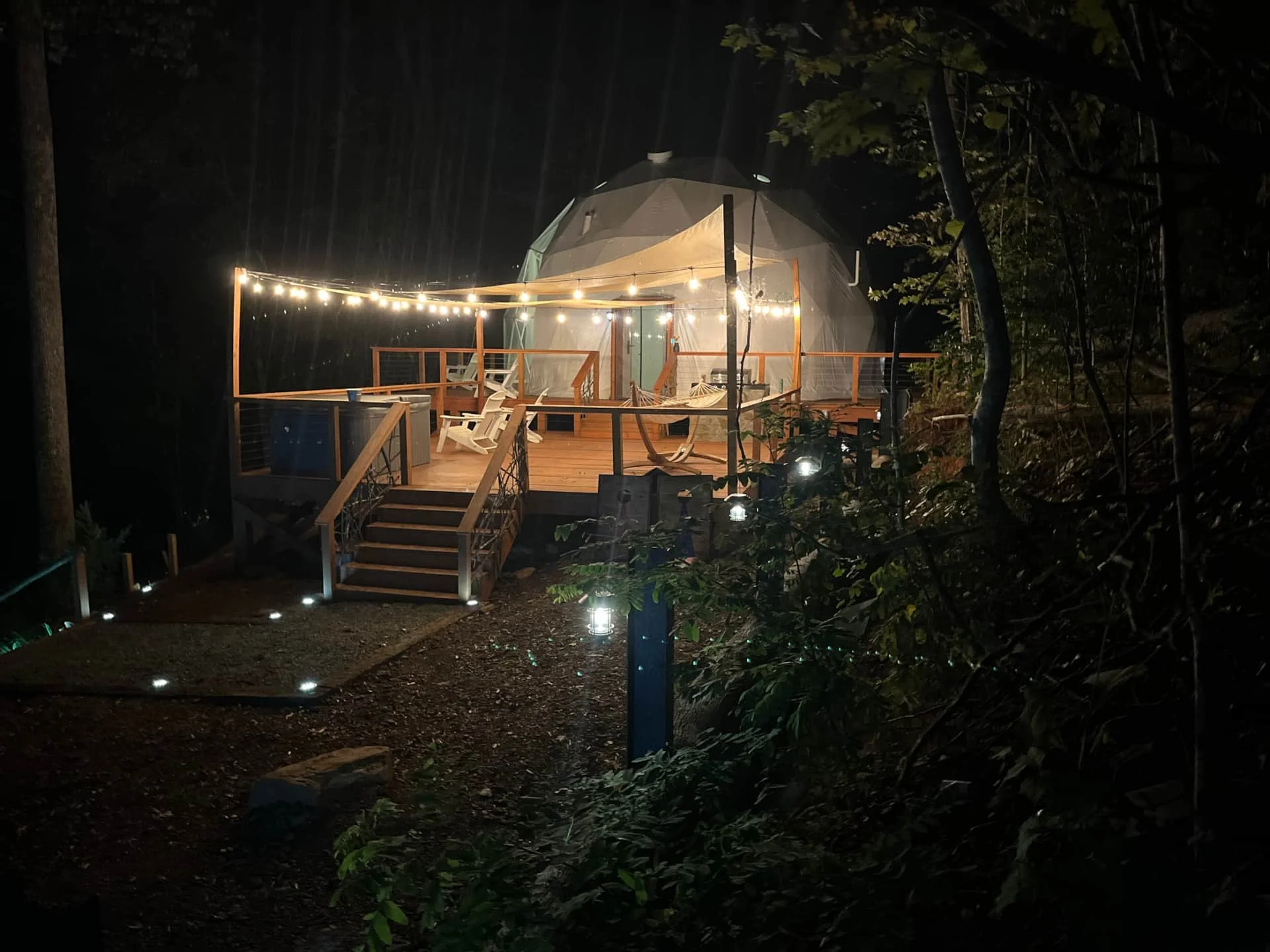 A lit, domed treehouse on a wooden deck with string lights at night in a forested area.