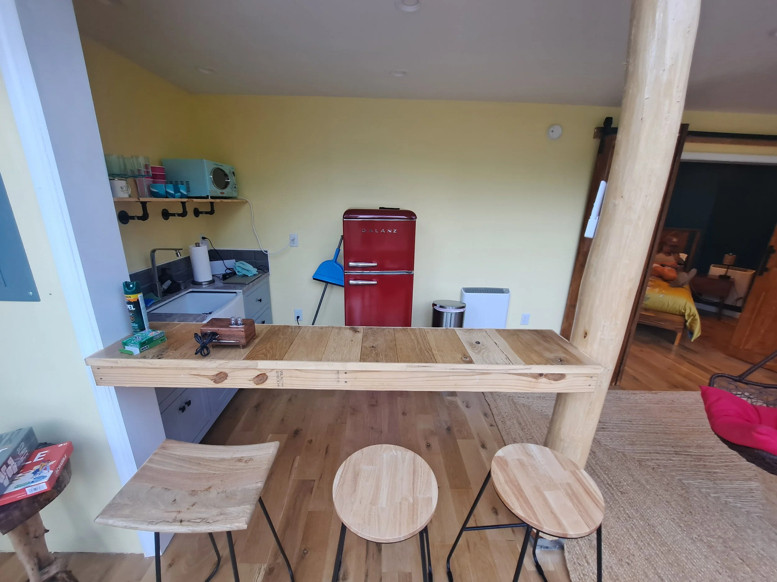 Kitchen with wooden countertops, a vintage red refrigerator, small bar stools, and a yellow wall, visible through an open wall with a wooden counter.
