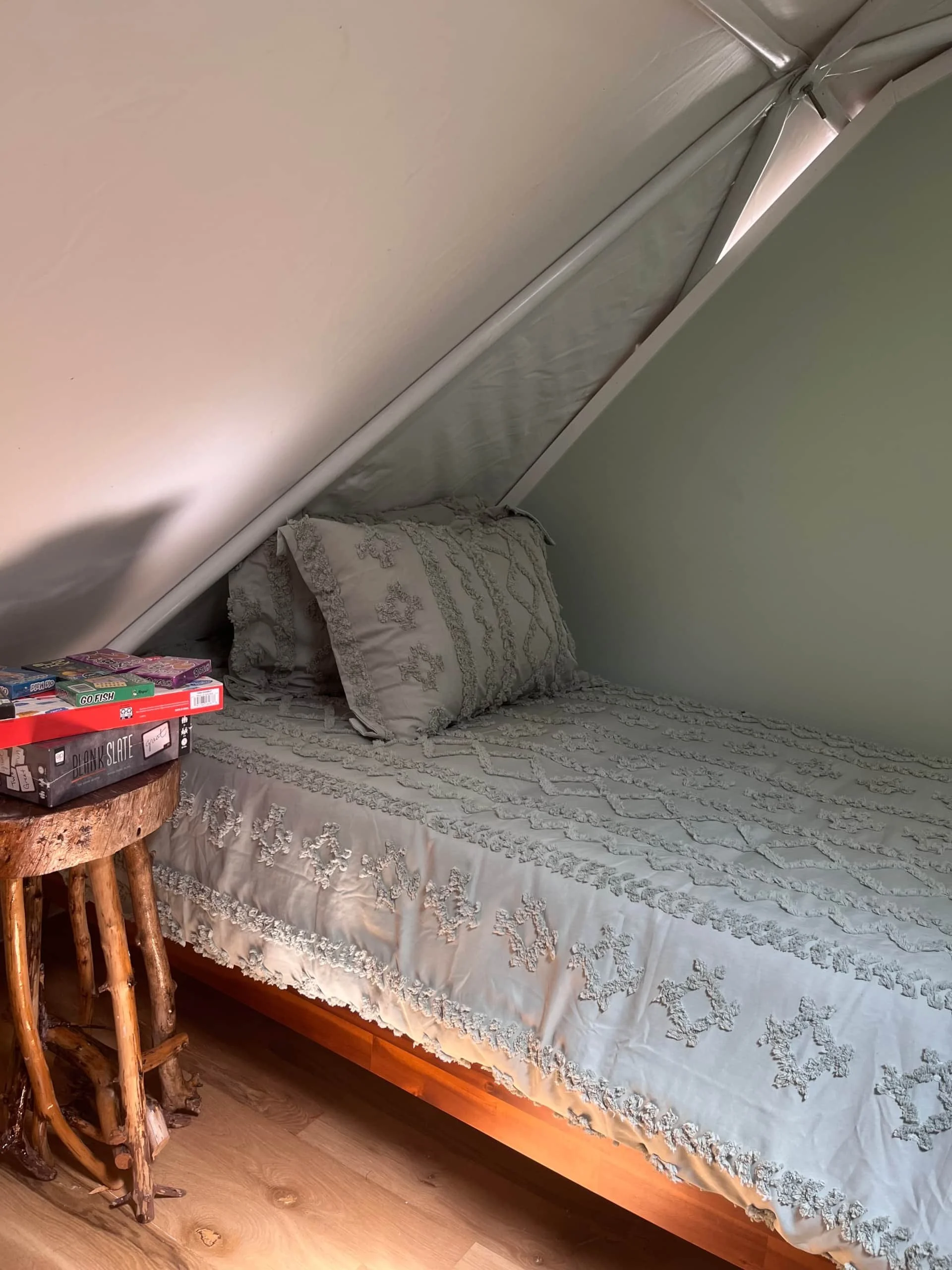 A bedroom with an angled ceiling, a bed with a embroidered bedspread and matching pillow, and a small rustic wooden side table with board games on top.