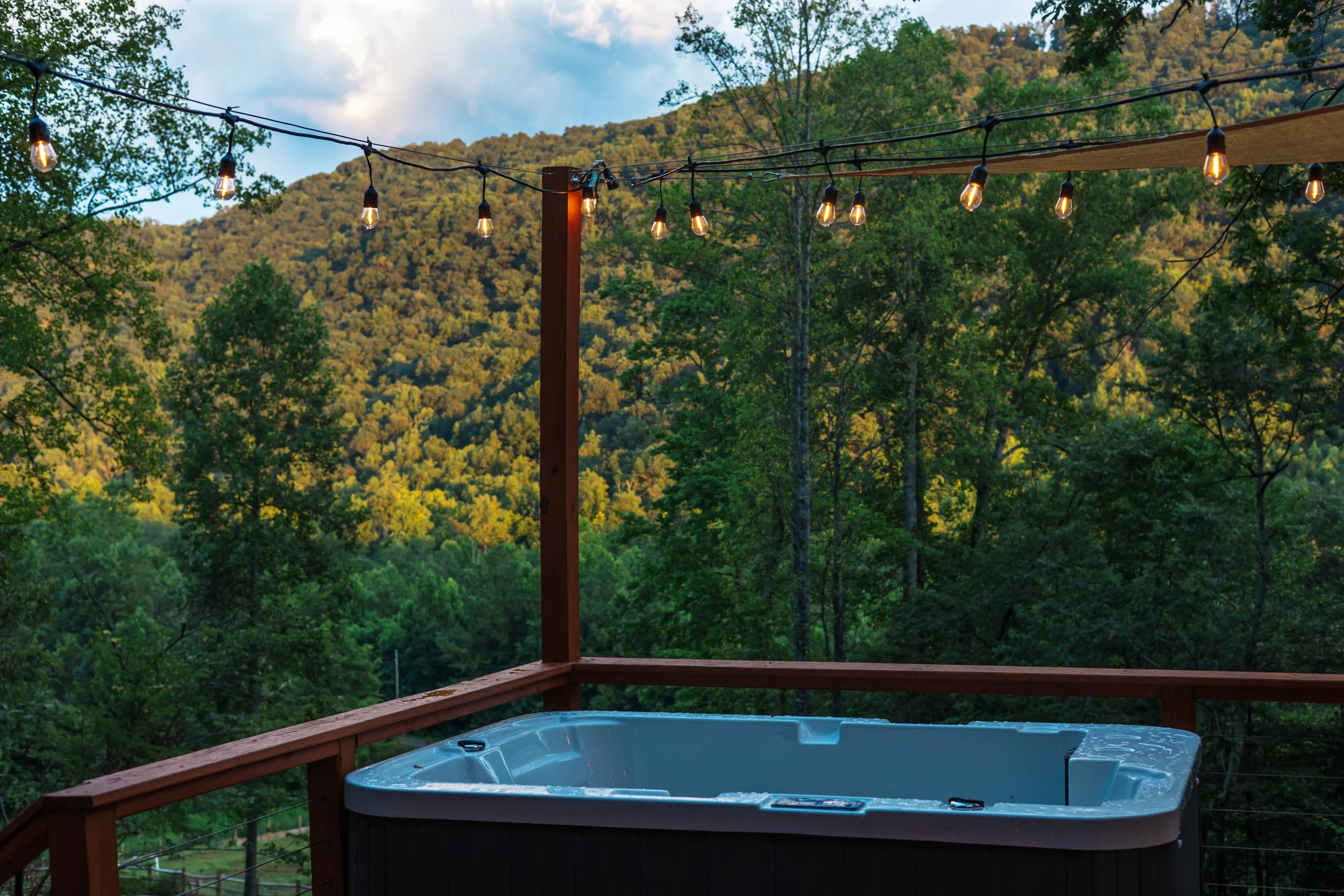 Hot tub on a wooden deck overlooking a green forested mountain landscape with string lights overhead.