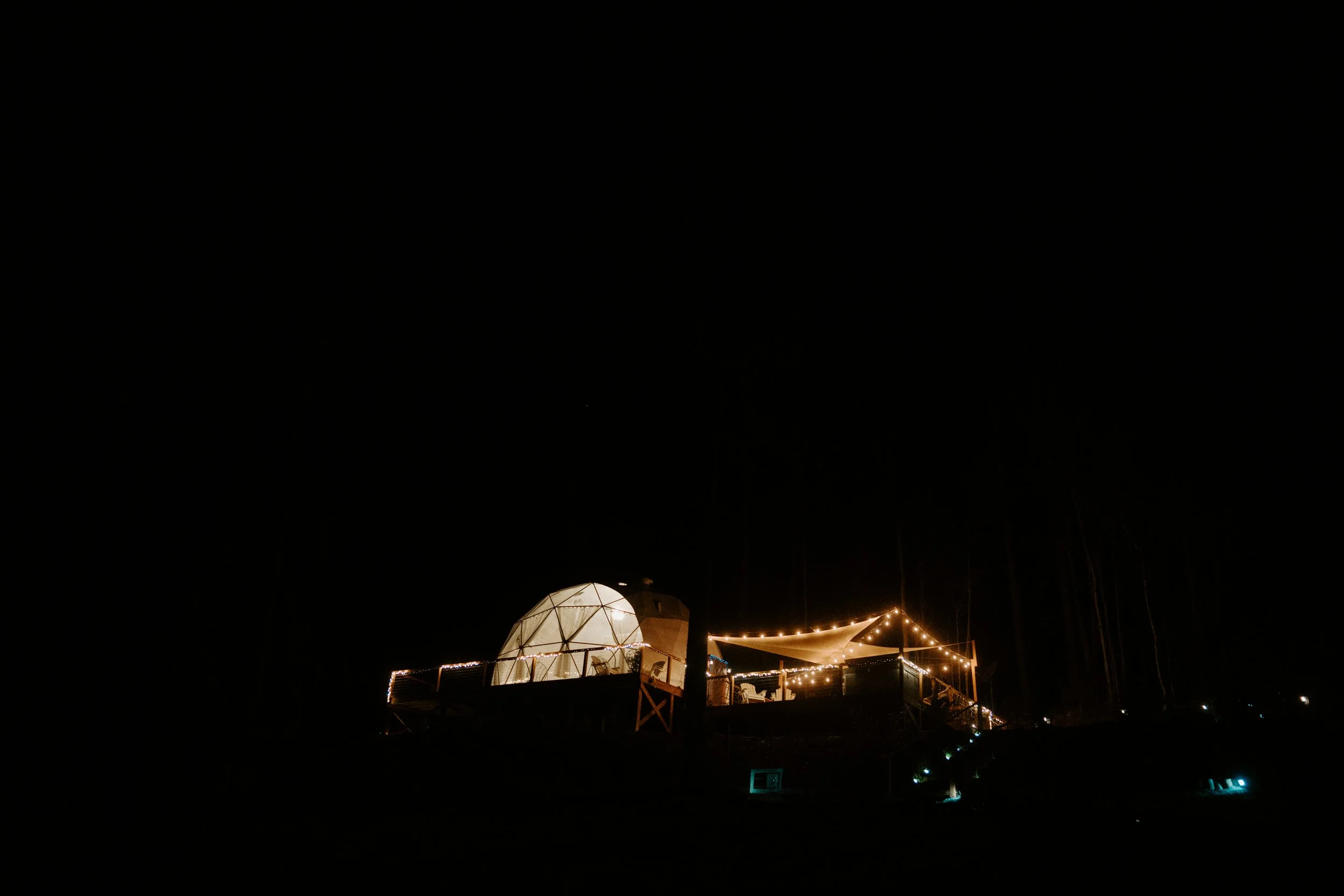 Nighttime scene of a lit structure with a geodesic dome, string lights, and a canopy on a dark background.