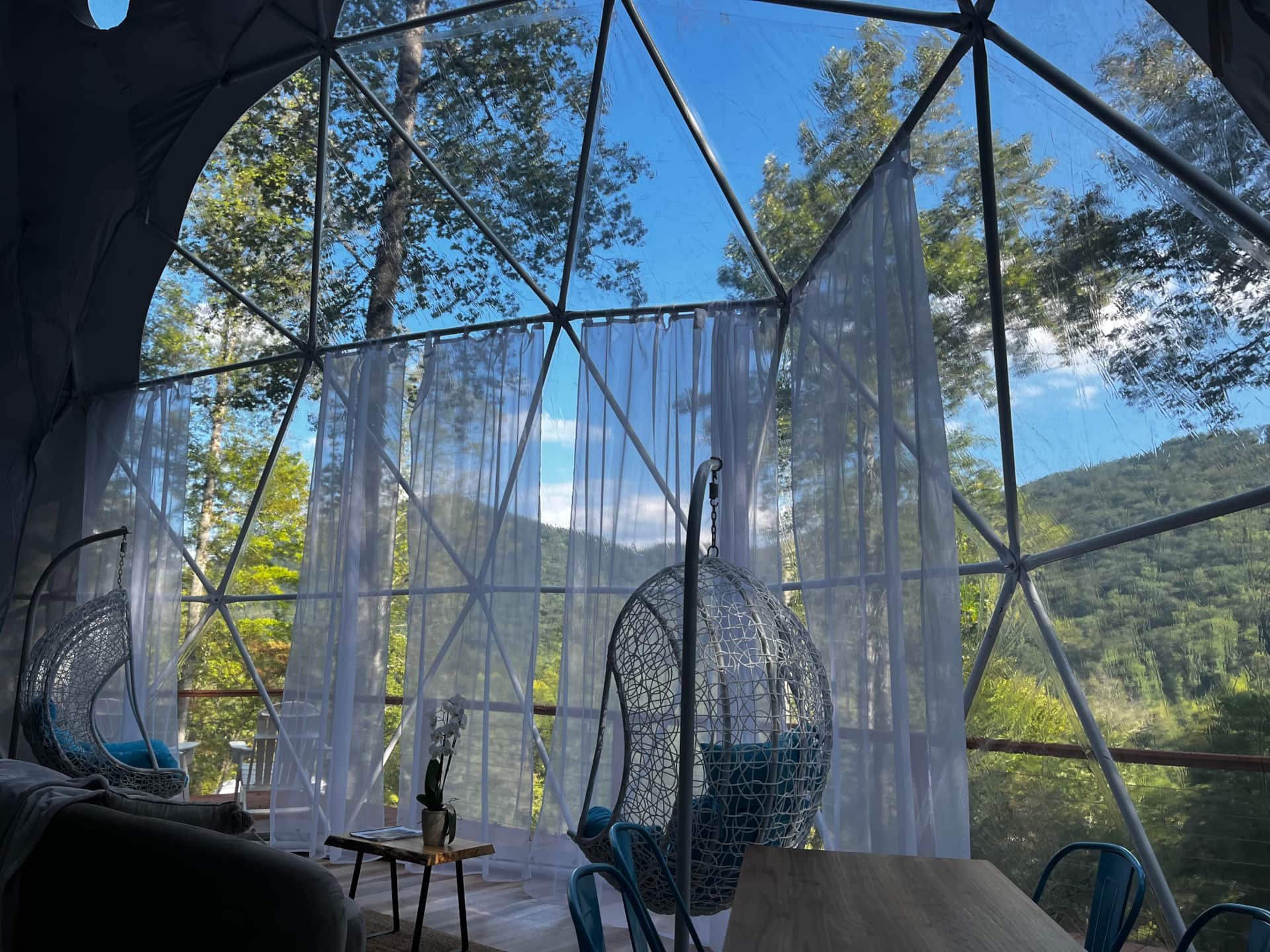 Interior of a geodesic dome with large windows, white curtains, hanging wicker chairs, a small table with a potted orchid, and blue chairs, overlooking a forested mountain landscape under a clear blue sky.